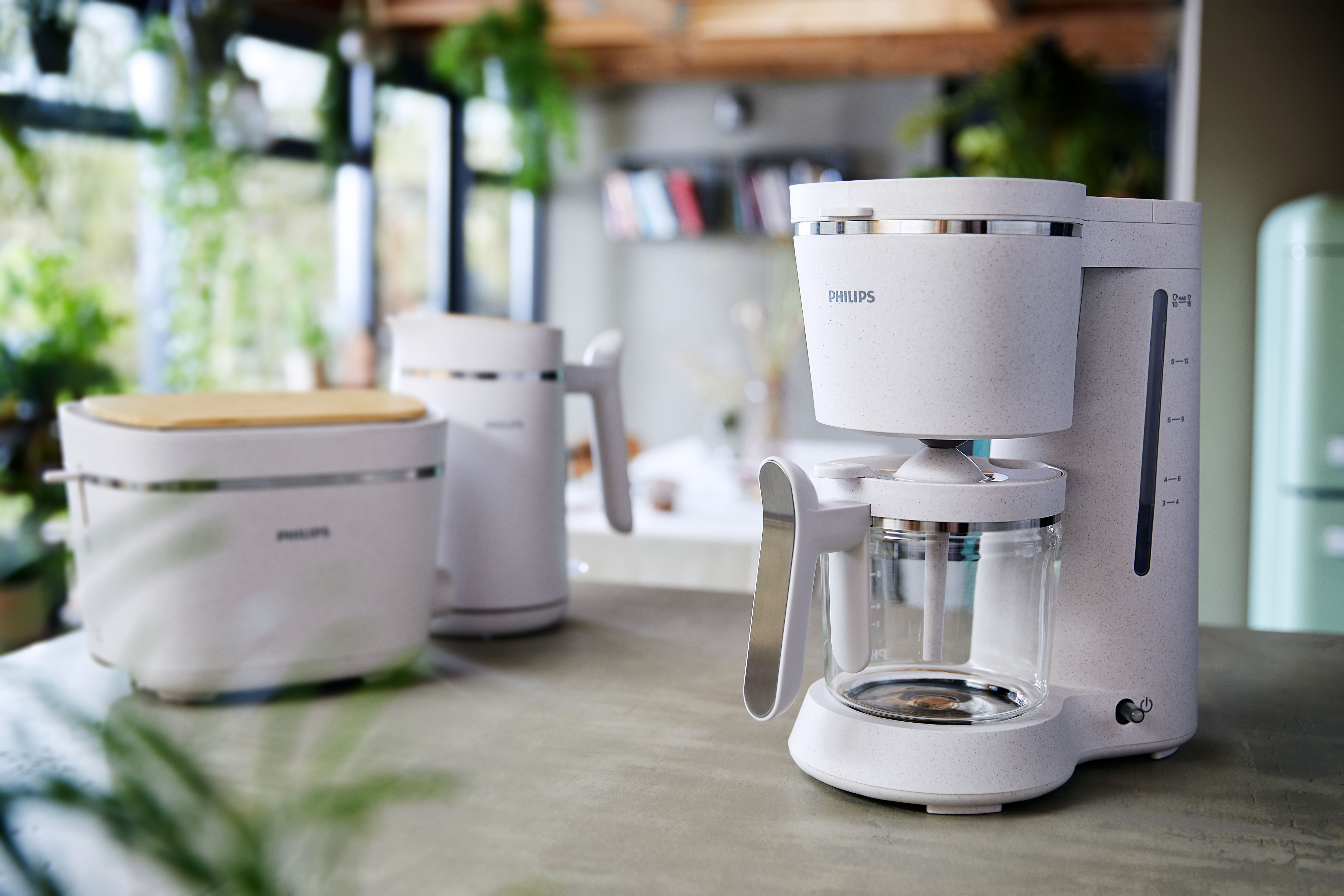 White coffee machine and accessories. Wooden counter. Green plants blurred in the background.