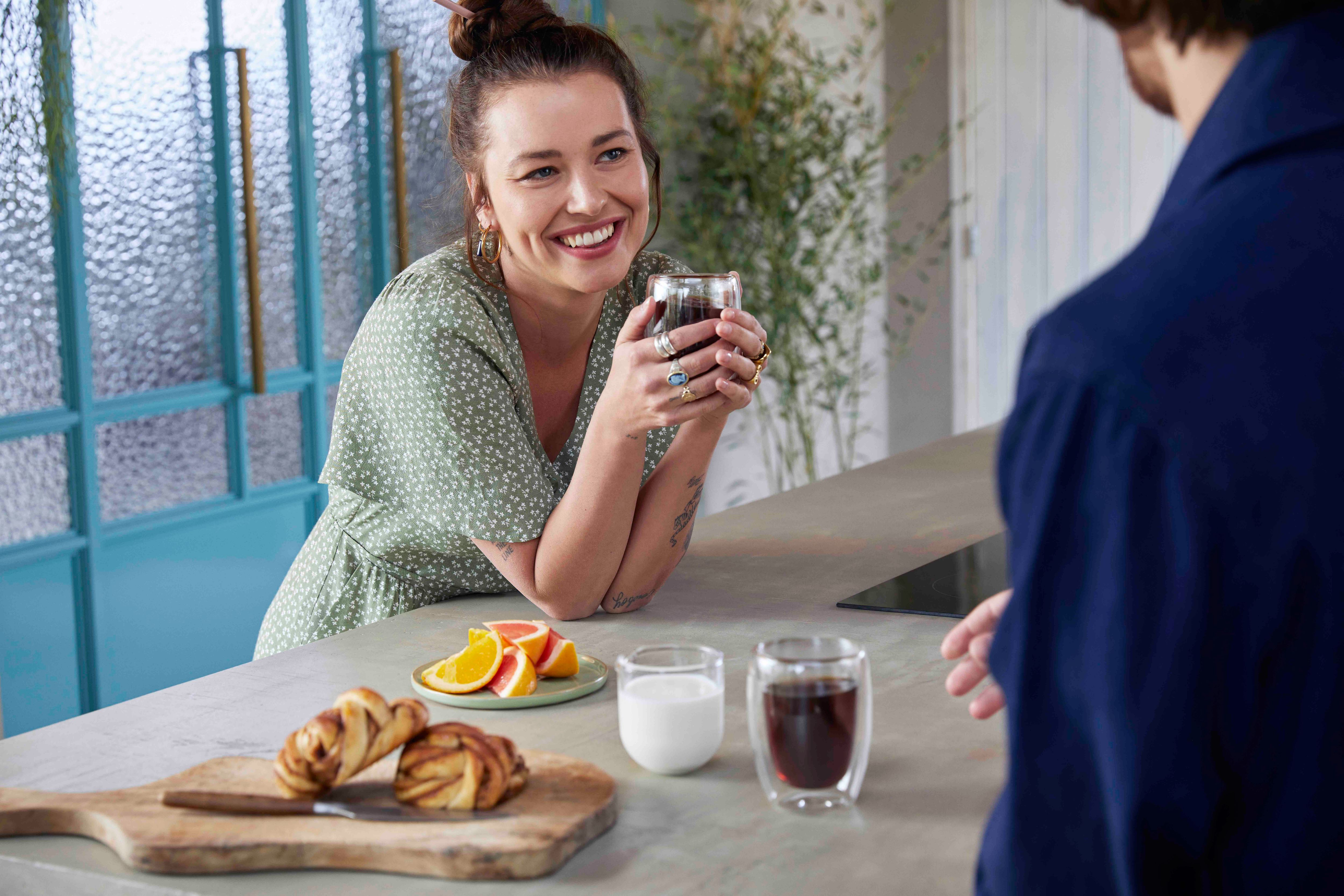 Woman smiling, holding coffee cup, with a man in the background. Food on the table.
