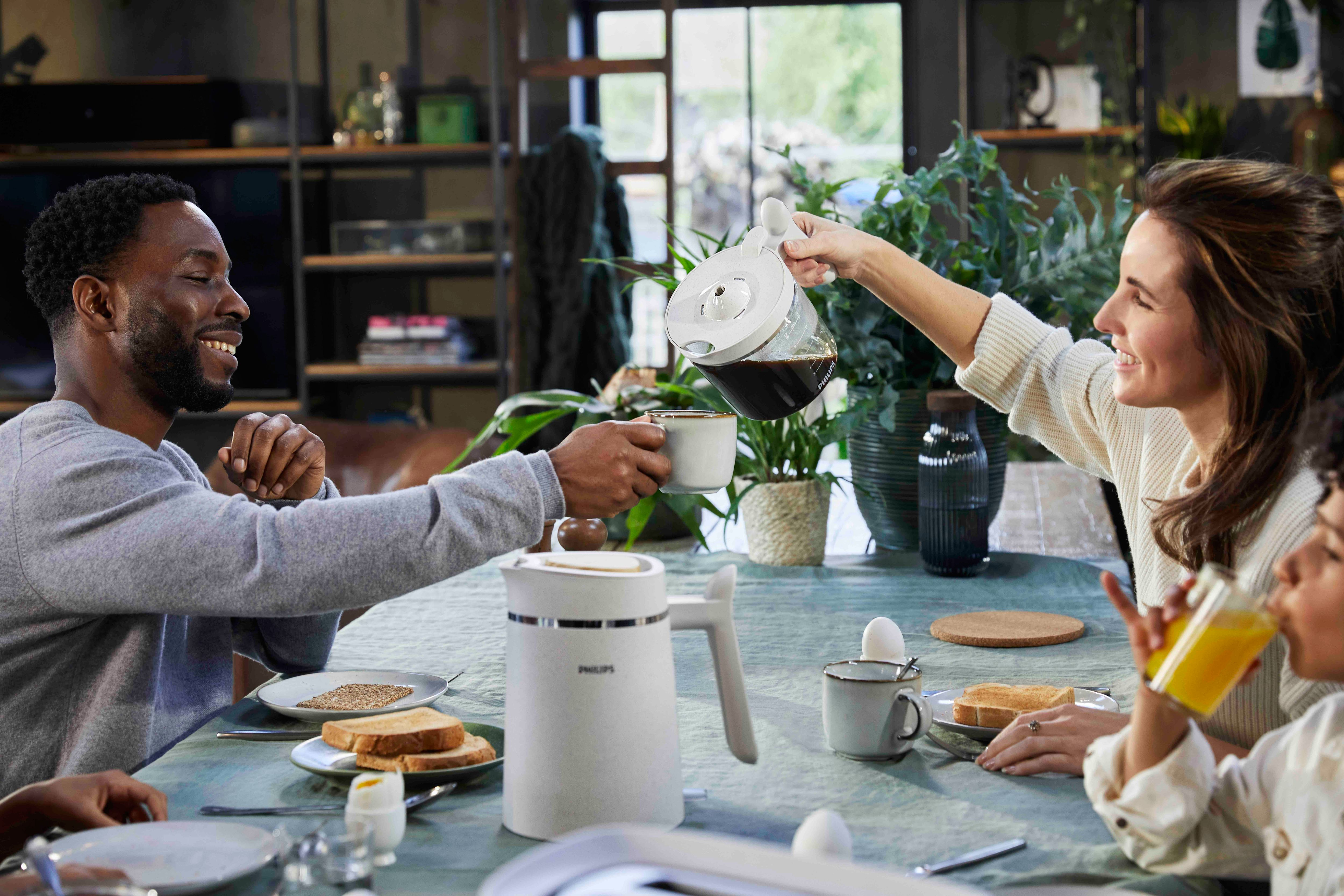 Family at breakfast, woman pouring coffee. Table set with food and coffee maker.