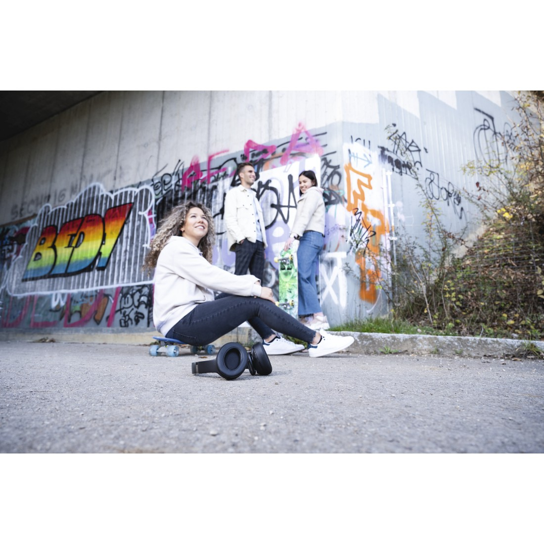 Drei Personen sitzen und stehen in der Nähe einer Graffiti-Wand mit Skateboards und Kopfhörern.