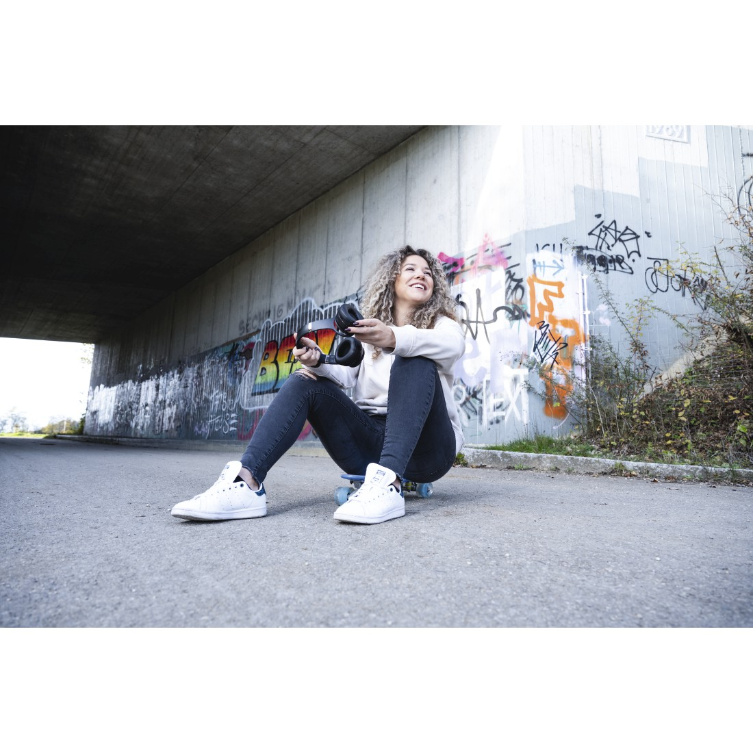 Frau auf Skateboard unter Brücke, Kopfhörer haltend, lächelnd.