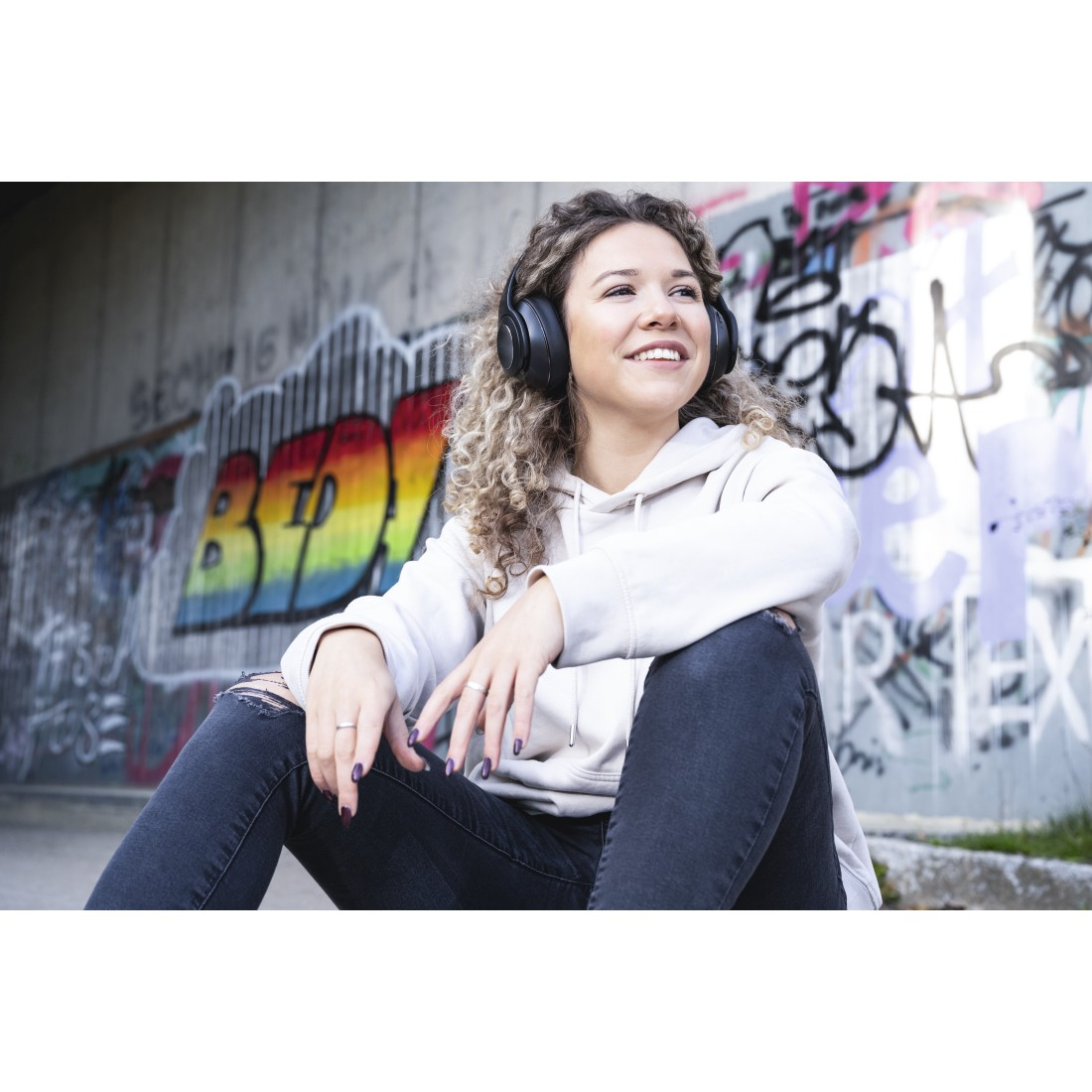 Eine Frau mit Kopfhörern sitzt vor Graffiti. Sie lächelt und blickt nach oben.