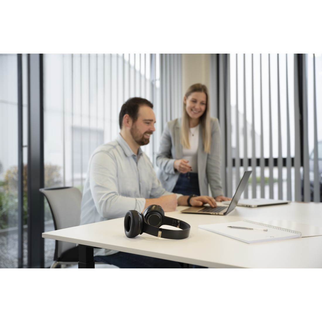 Zwei Personen, ein Mann und eine Frau, sitzen an einem weißen Tisch mit einem Laptop und Kopfhörern. Sie lächeln.