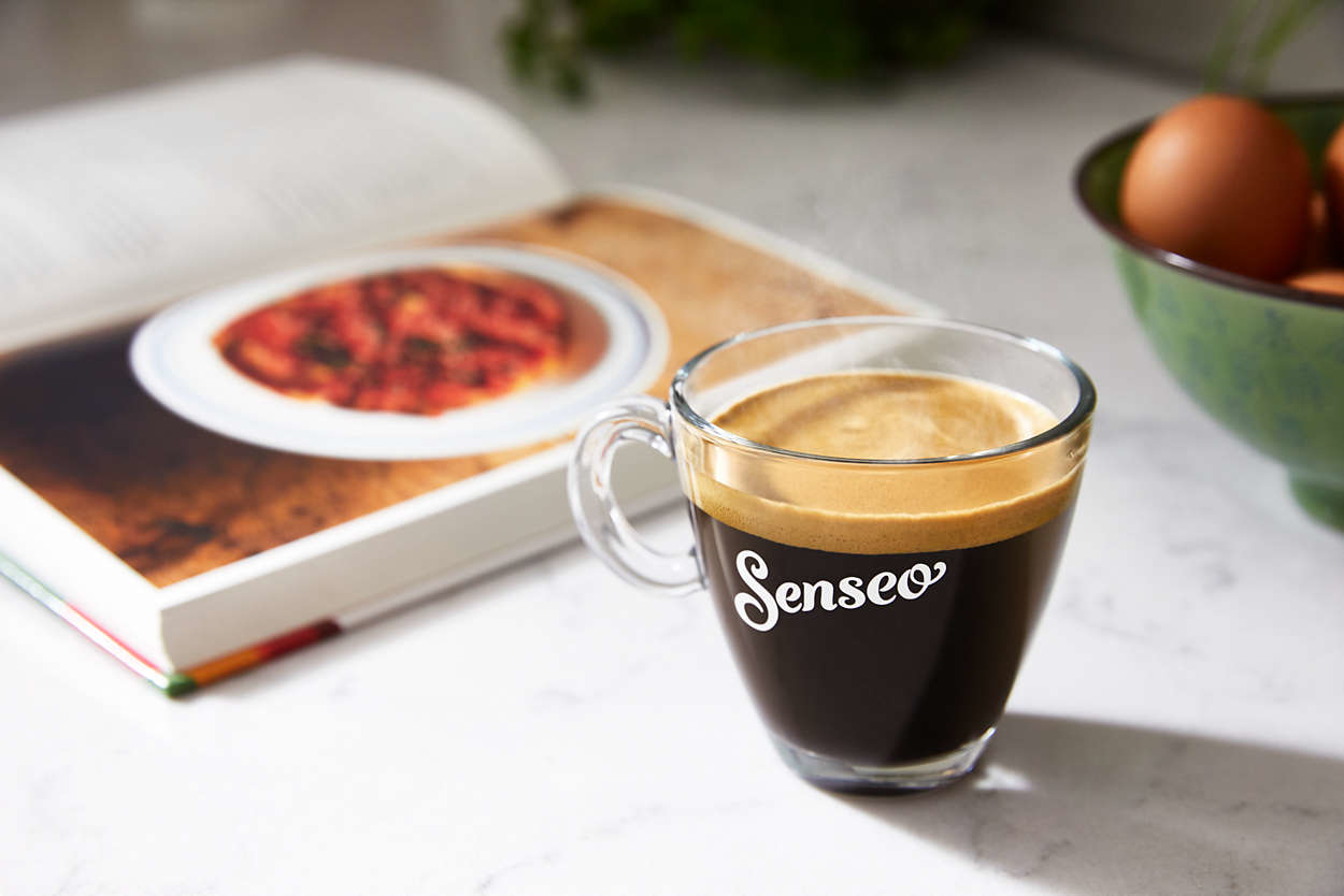 Eine Tasse Kaffee mit dem Wort 'Senseo' darauf, die auf einem Tisch mit einem Kochbuch steht.