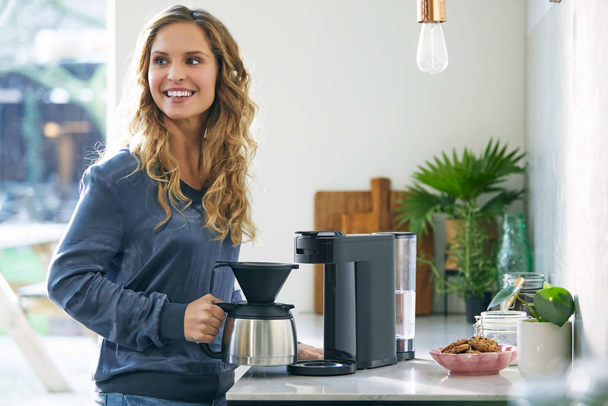 Een vrouw glimlacht terwijl ze een koffiepot vasthoudt naast een zwarte koffiemachine.