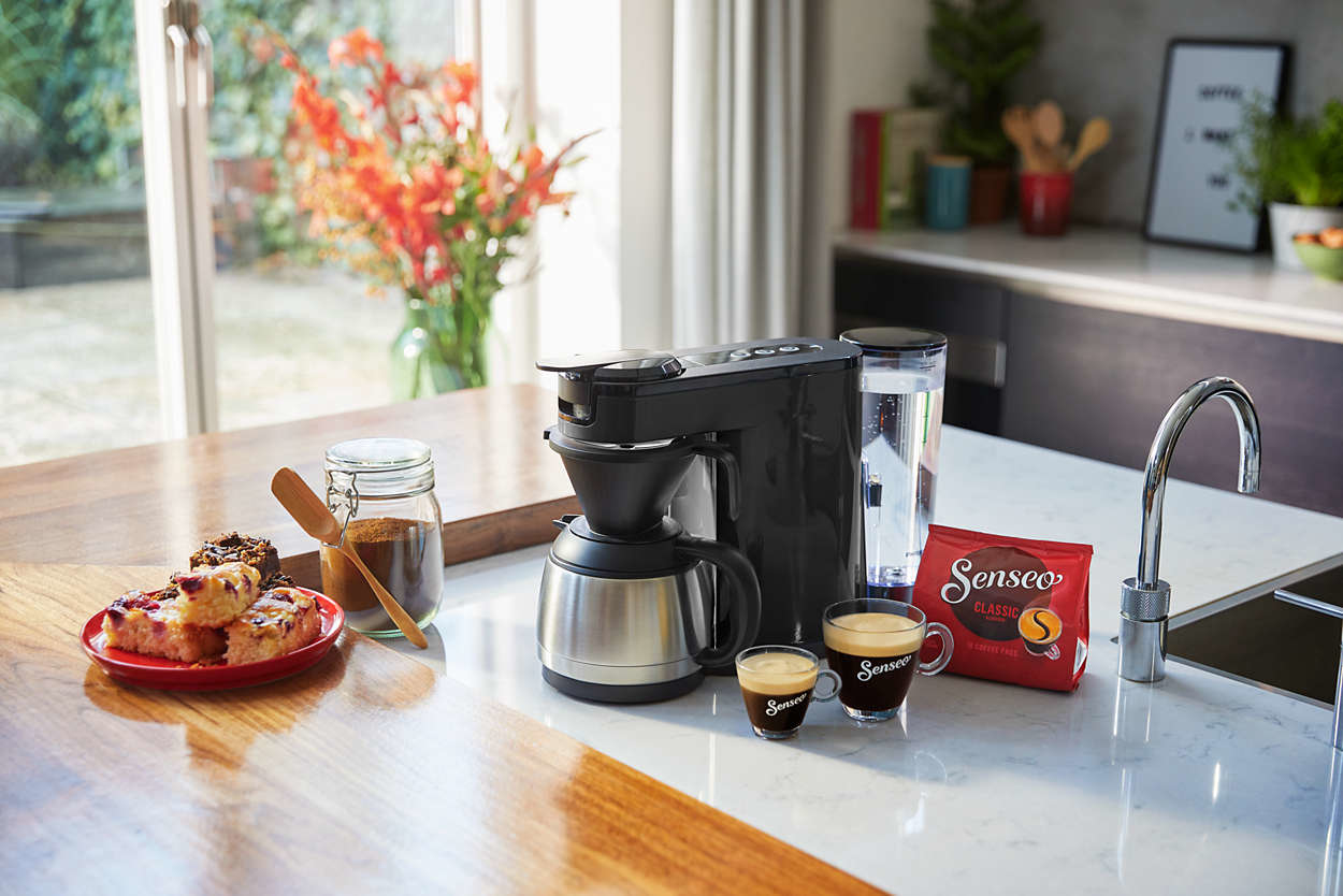 Een koffiezetapparaat met kopjes, een koffieverpakking, gebak en bloemen op een wit aanrecht.