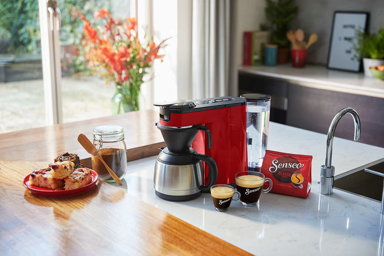 Een rode Senseo-koffiemachine met koffie, lekkernijen en accessoires op een aanrecht.
