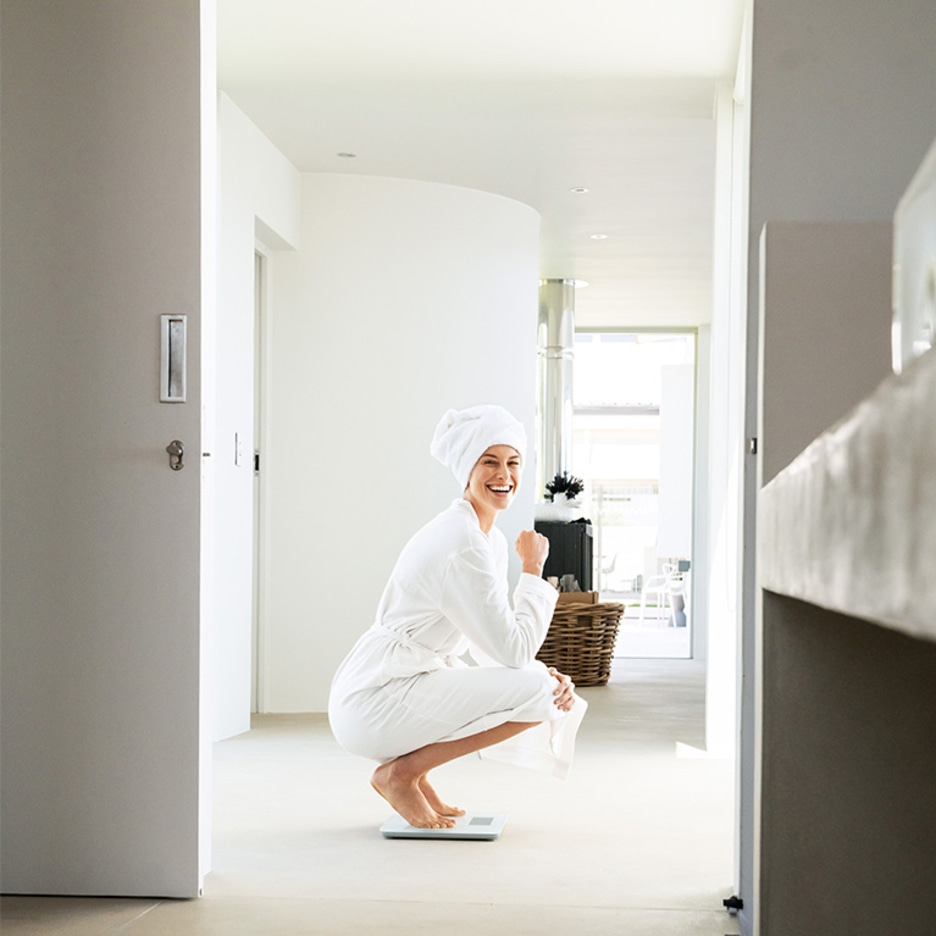 Femme en peignoir sur balance, souriante, salle de bain. Couloir blanc lumineux avec porte et lavabo.
