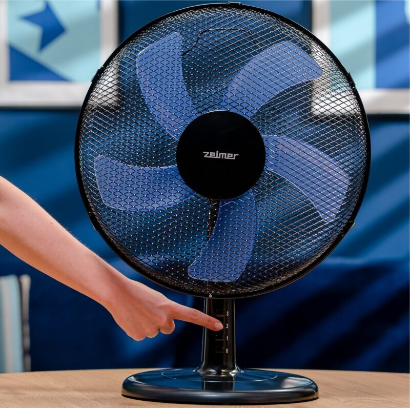 Czarny wentylator z niebieskimi łopatkami, ręka wskazująca ustawienie prędkości na stole.