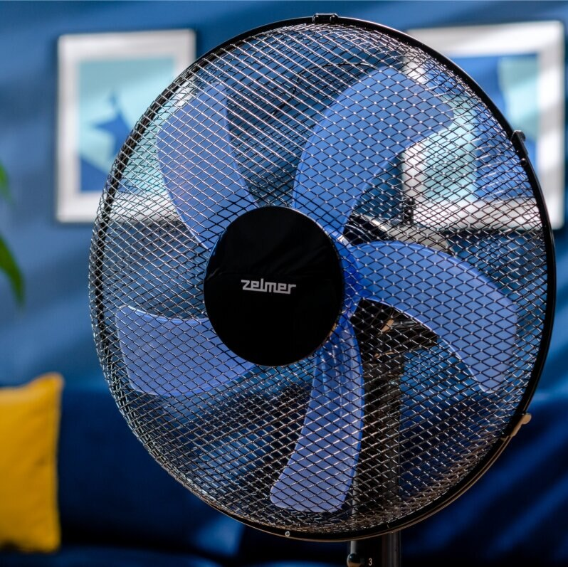 Zbliżenie niebieskiego wentylatora z czarną ramą. Tło stanowią niebieskie ściany i sofa.