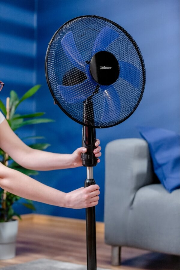 Kobieta w kuchni, trzymająca czarno-niebieski wentylator podłogowy Zelmer, rozmyte tło.