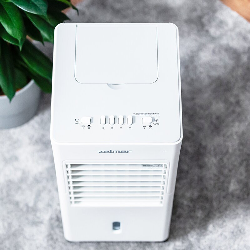 Biała chłodziarka powietrza z przyciskami sterowania. Stoi na szarej powierzchni, z rośliną w tle.