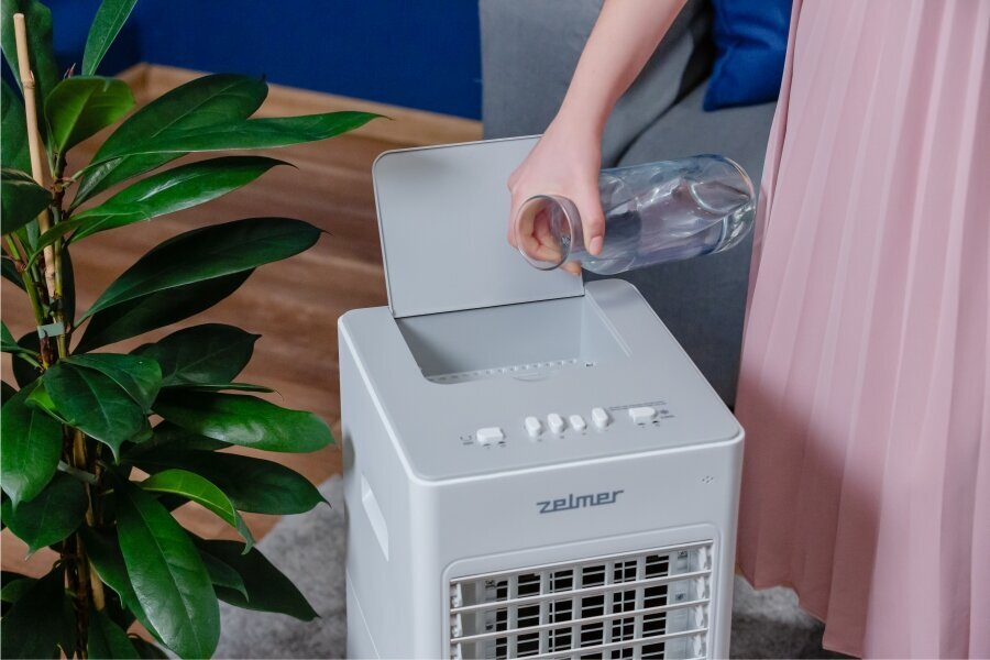 Kobieta wlewa wodę do białej chłodziarki powietrza, obok roślina.