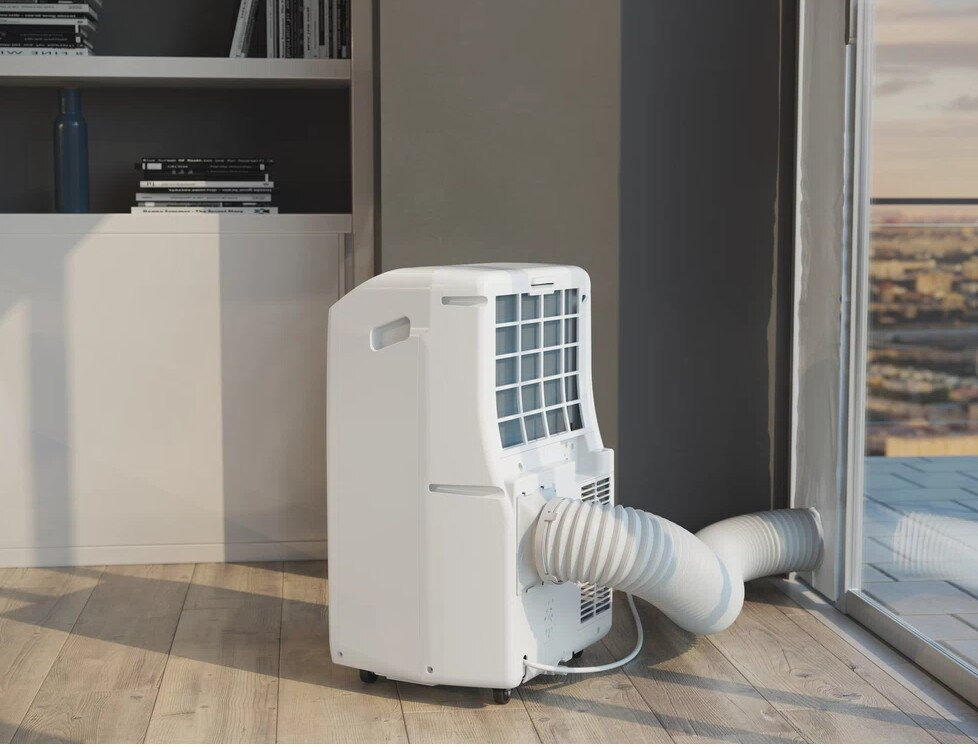 Biały przenośny klimatyzator stoi blisko otwartego okna. Elastyczna rura wystaje na zewnątrz. Regał na książki jest w tle.