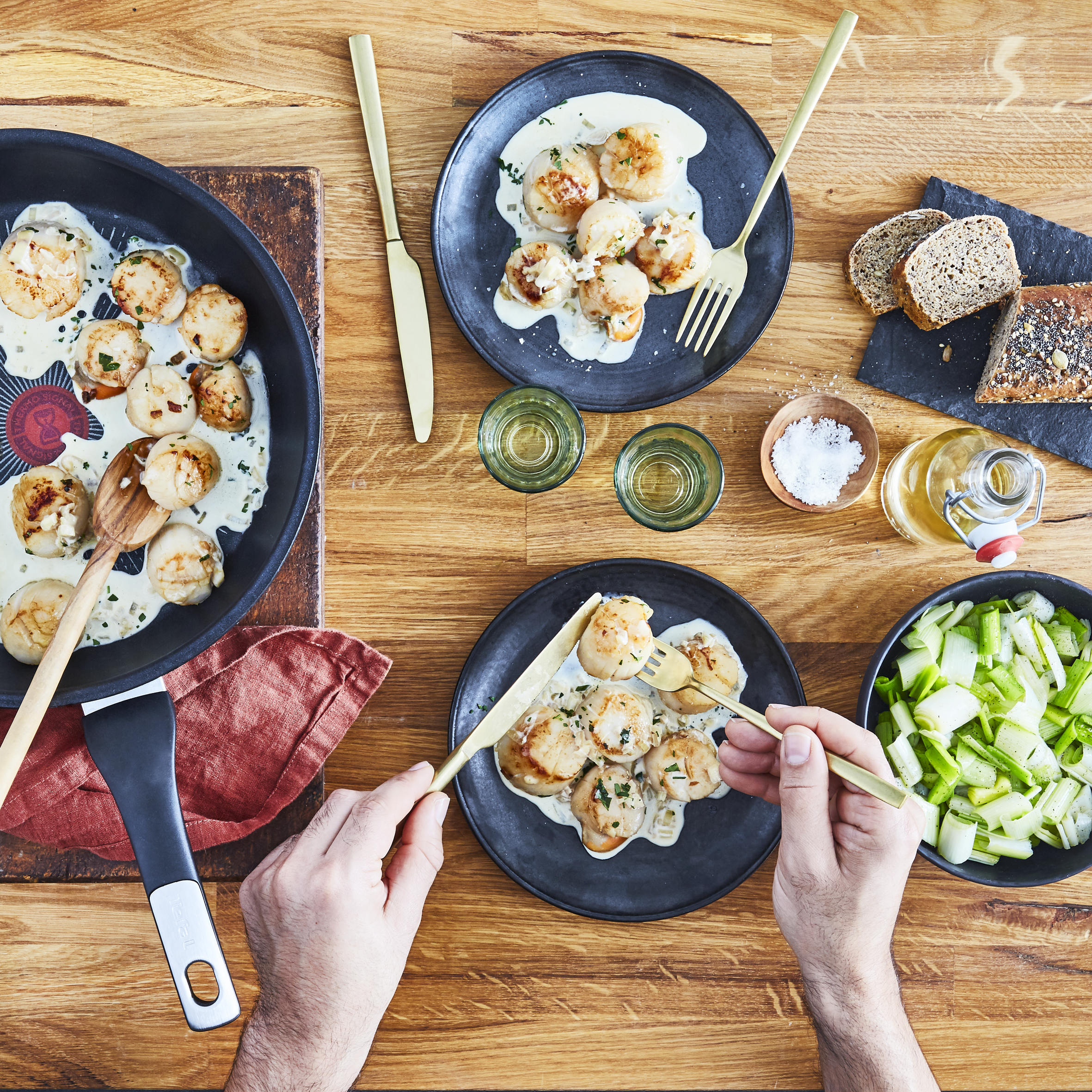 Vieiras en una sartén, en platos y gente comiendo. Mesa de madera con platos de comida.