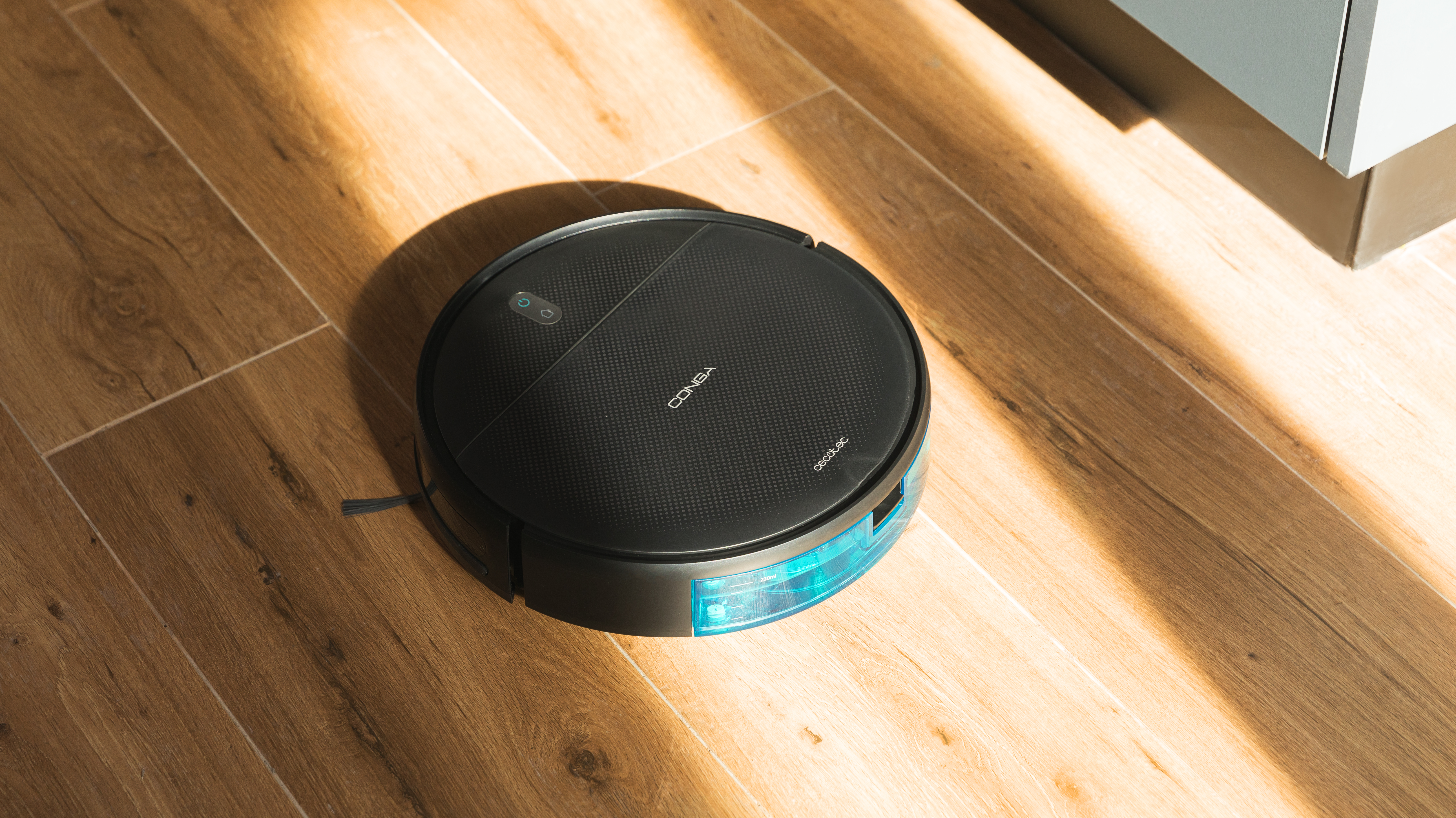 Aspiradora robot negra en un suelo de madera, con luz solar y un armario a la vista.