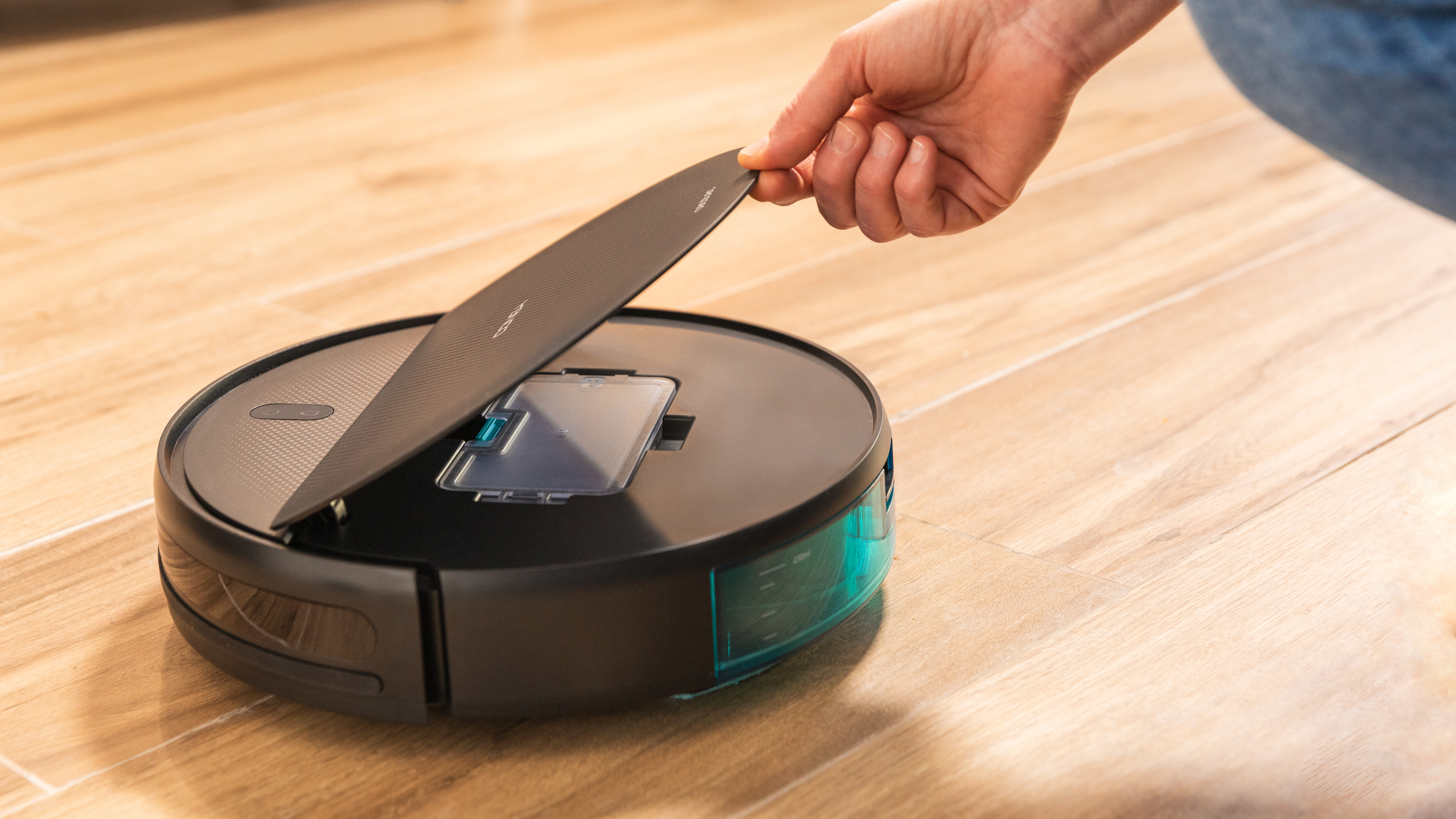 Mano levantando la tapa de una aspiradora robot negra en suelo de madera.