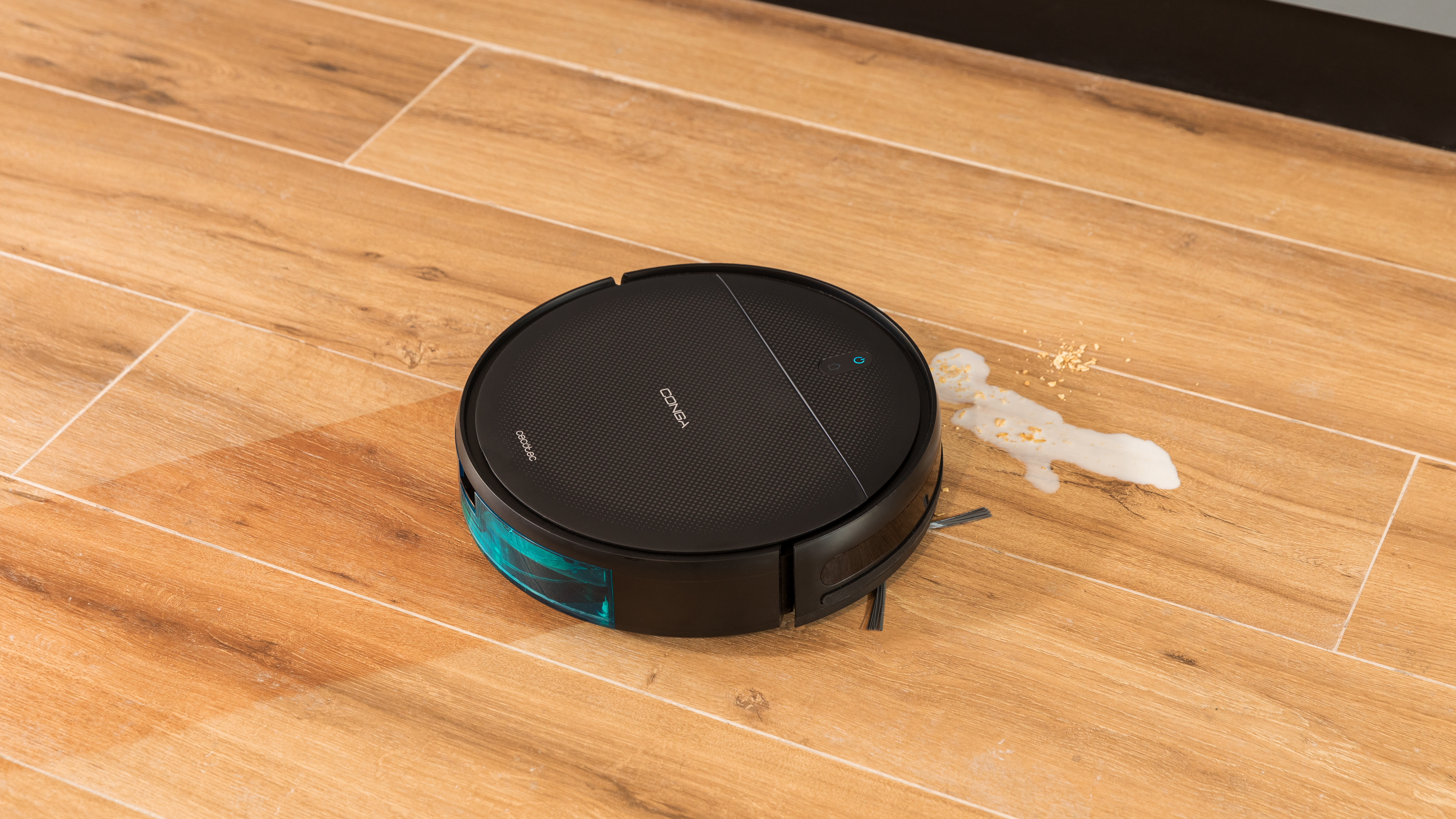 Una aspiradora robot negra en un piso de madera con un derrame cerca.