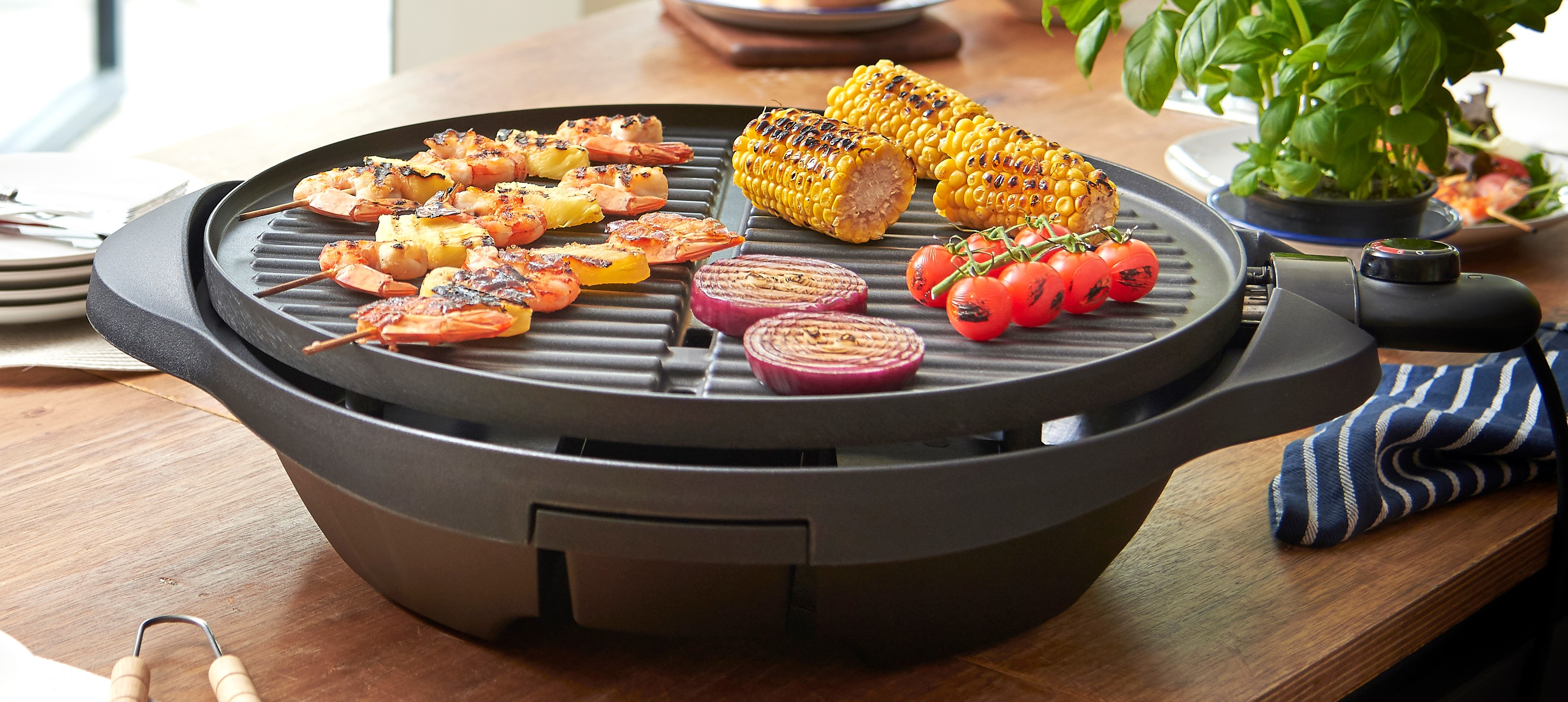 Gegrilltes Essen, darunter Garnelen, Mais und Zwiebeln, auf einem schwarzen Grill. Es steht auf einem Holztisch.