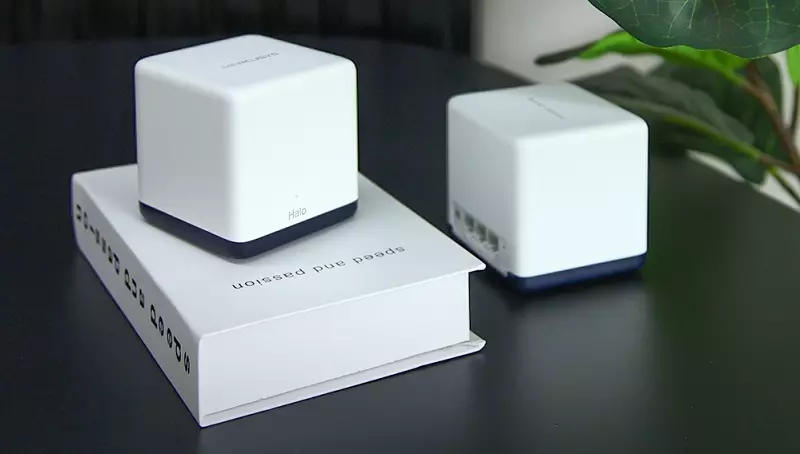 Dos dispositivos blancos en forma de cubo, uno sobre un libro, otro sobre una mesa negra. Planta verde al fondo.