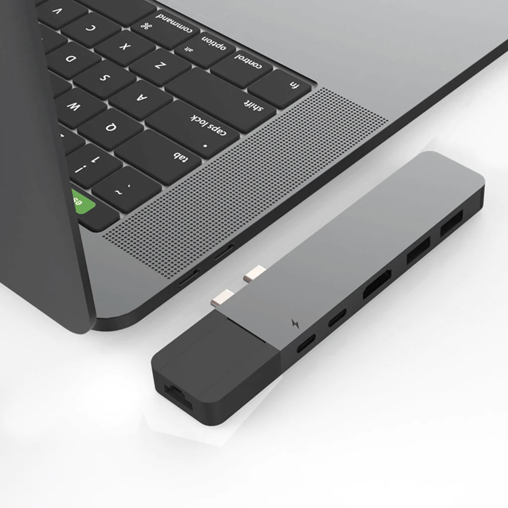 Un portátil conectado a un concentrador USB-C gris y negro con varios puertos, sobre una superficie blanca.