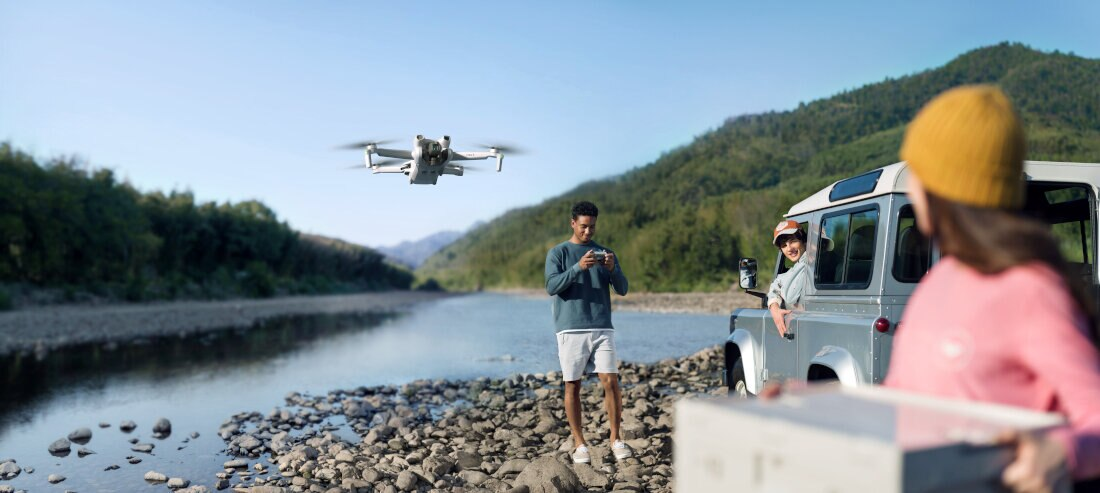 Dron lecący nad rzeką z ludźmi w pobliżu samochodu i górami w tle.