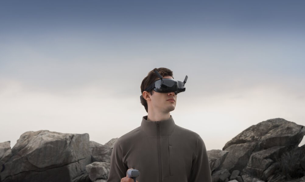 Mężczyzna w goglach VR, stojący na skałach, patrzący w górę, tło nieba.
