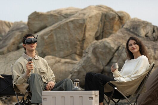 Dwie osoby na zewnątrz na krzesłach, jedna z goglami, druga z kubkiem, w pobliżu skał i stołu.