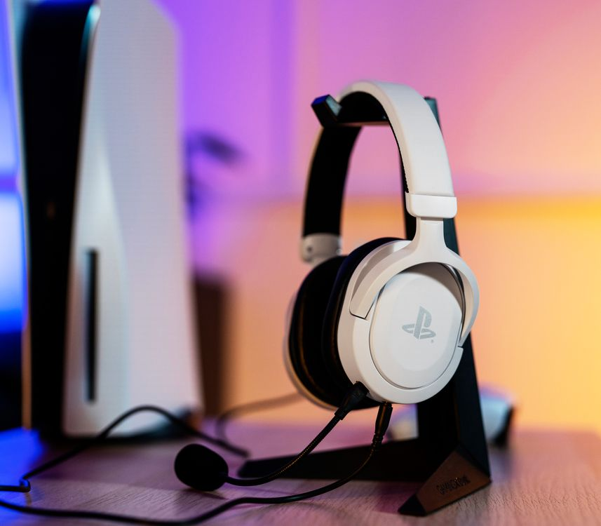 Fehér gamer headset egy állványon, fehér PS5 konzollal a háttérben.