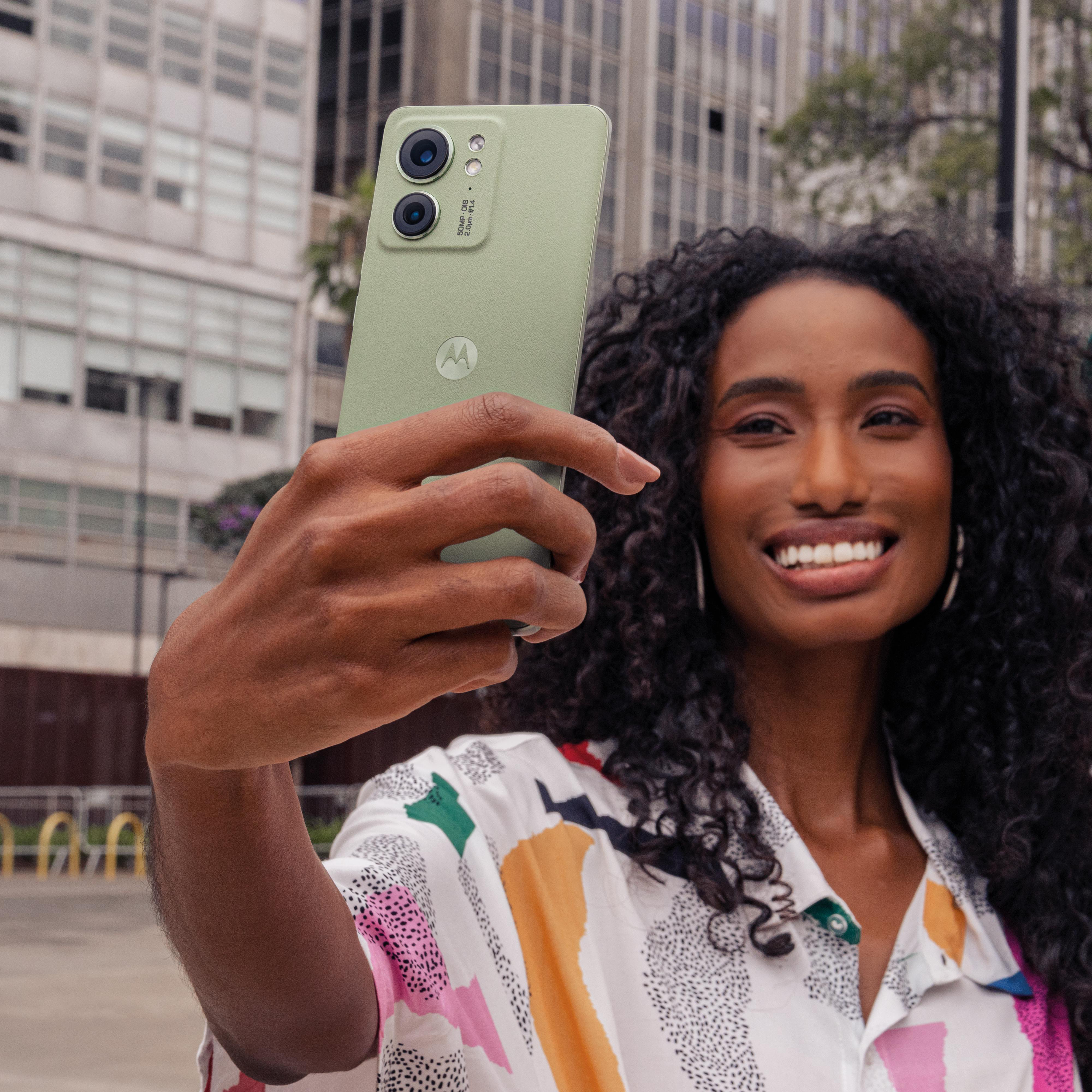 Frau macht Selfie mit grünem Telefon. Hintergrund ist eine Stadtstraße mit Gebäuden.
