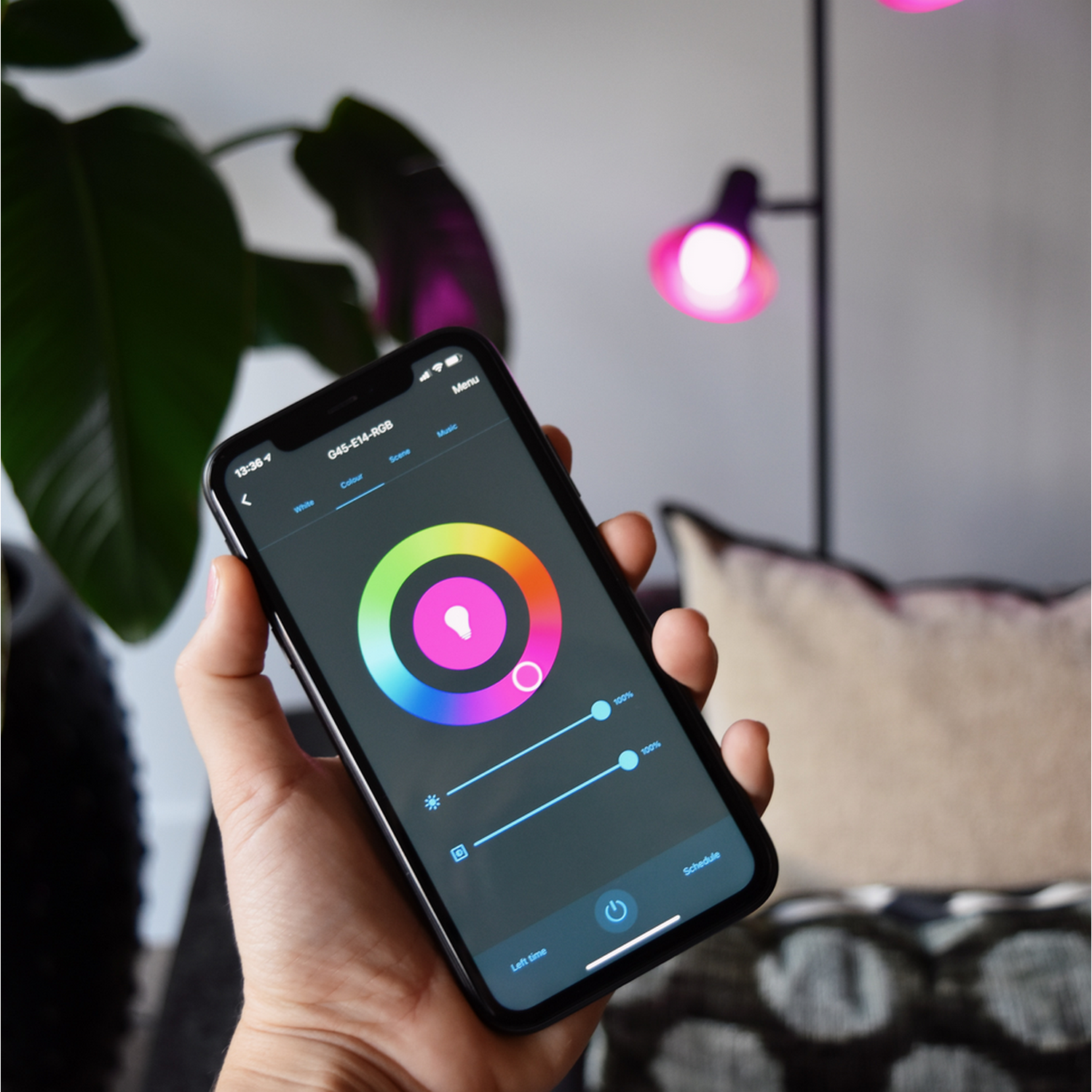 A hand holds a smartphone displaying a color wheel interface for lights. A lamp shines pink light. 