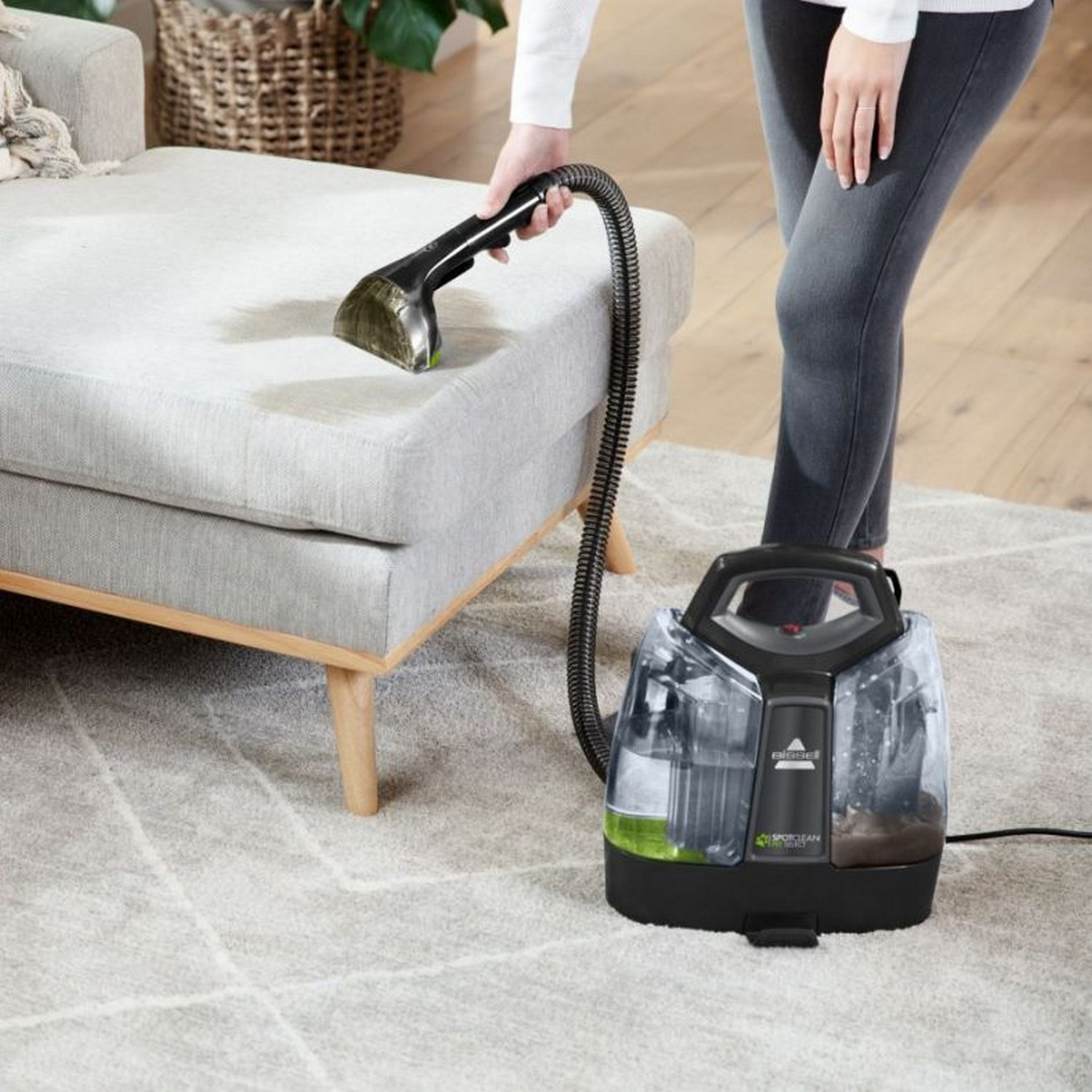 A person using a carpet cleaner on a grey sofa. The machine is on the carpet next to it.