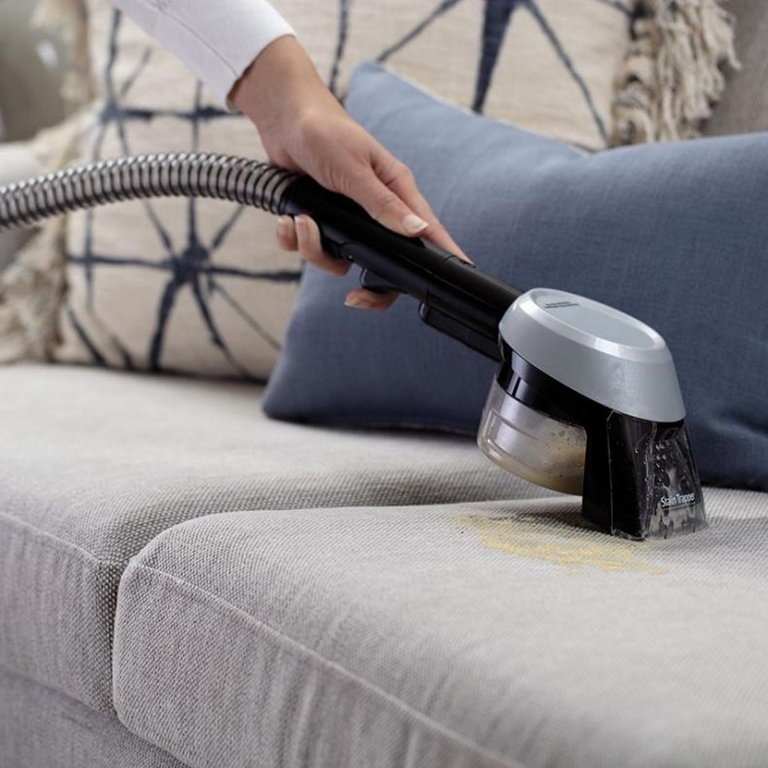 A person cleaning a sofa with a handheld cleaner, removing a stain. The sofa and cleaner are visible.
