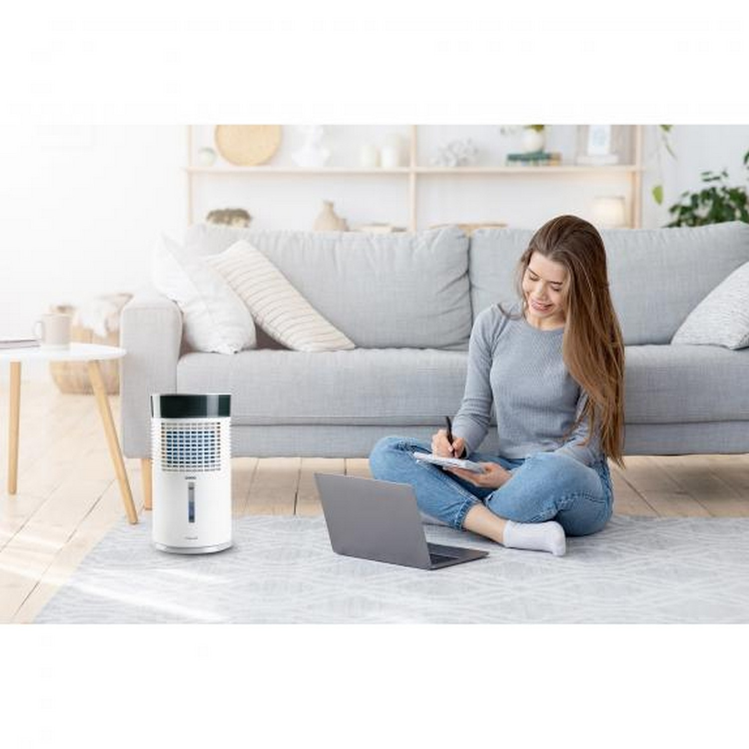 Une femme écrit sur un bloc-notes tout en étant assise par terre avec un ordinateur portable et un refroidisseur d'air.