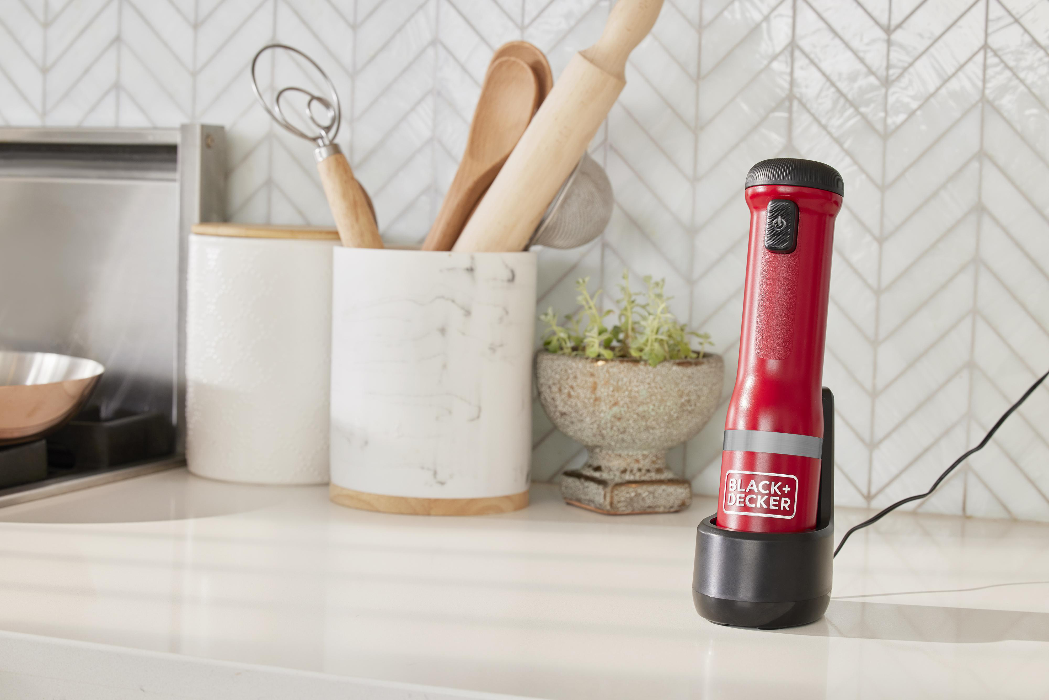Roter und schwarzer kabelloser Handmixer in einer Ladestation, mit Küchenutensilien im Hintergrund.
