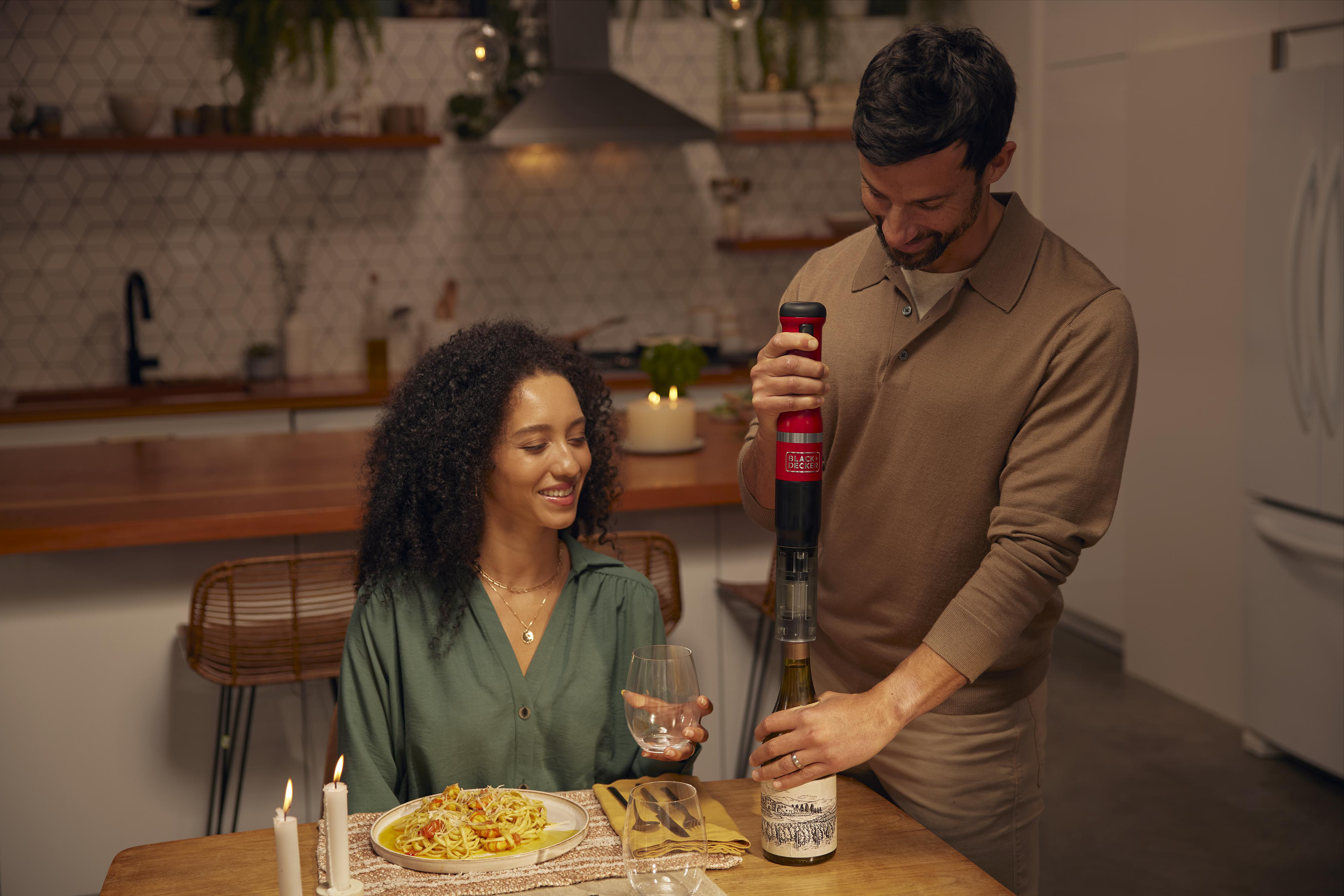 Ein Mann benutzt einen roten Weinöffner an einer Flasche, während eine Frau am Tisch zusieht.