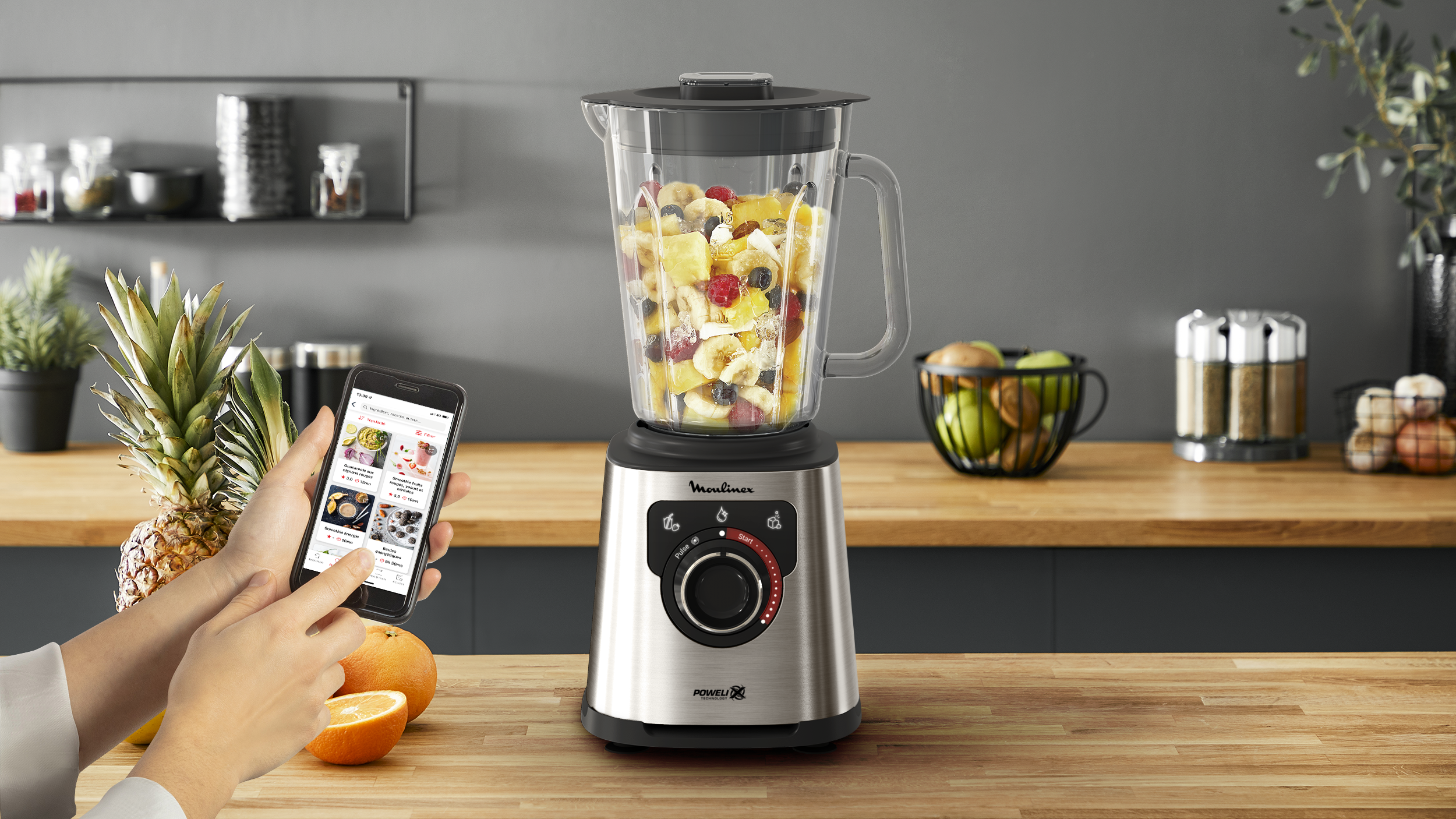 Ein Mixer mit Früchten. Die Hand einer Person benutzt ein Telefon mit Rezepten. Tisch mit Früchten und Gläsern.