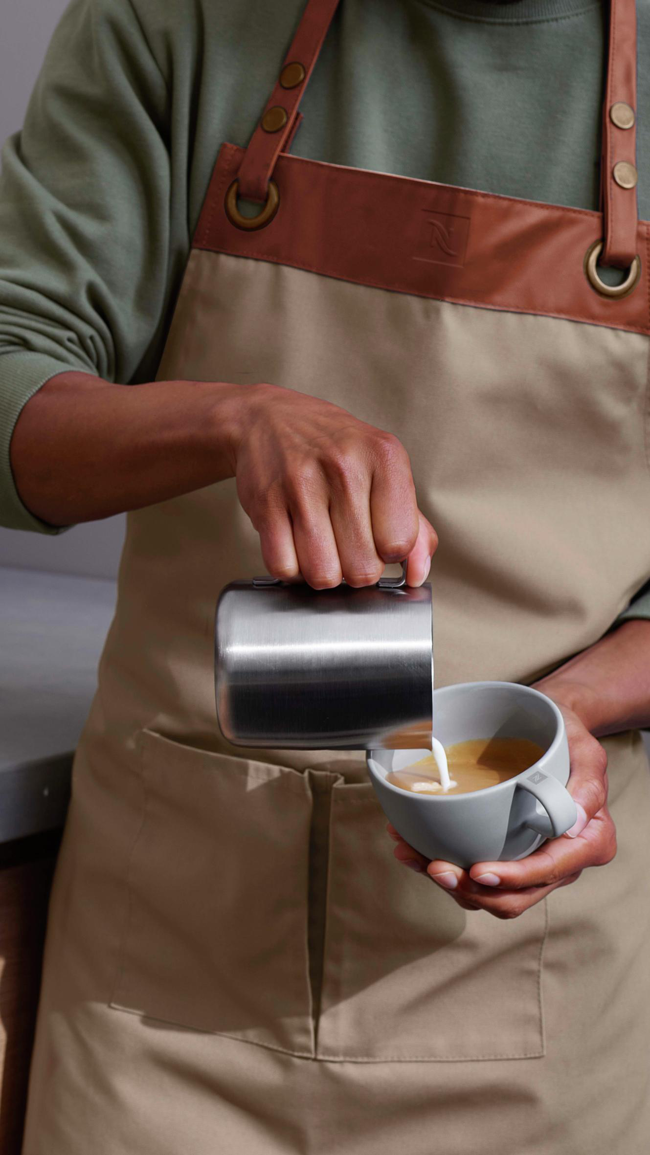 Eine Person, die Milch in eine Tasse Kaffee gießt, trägt eine beige Schürze, Nahaufnahme.
