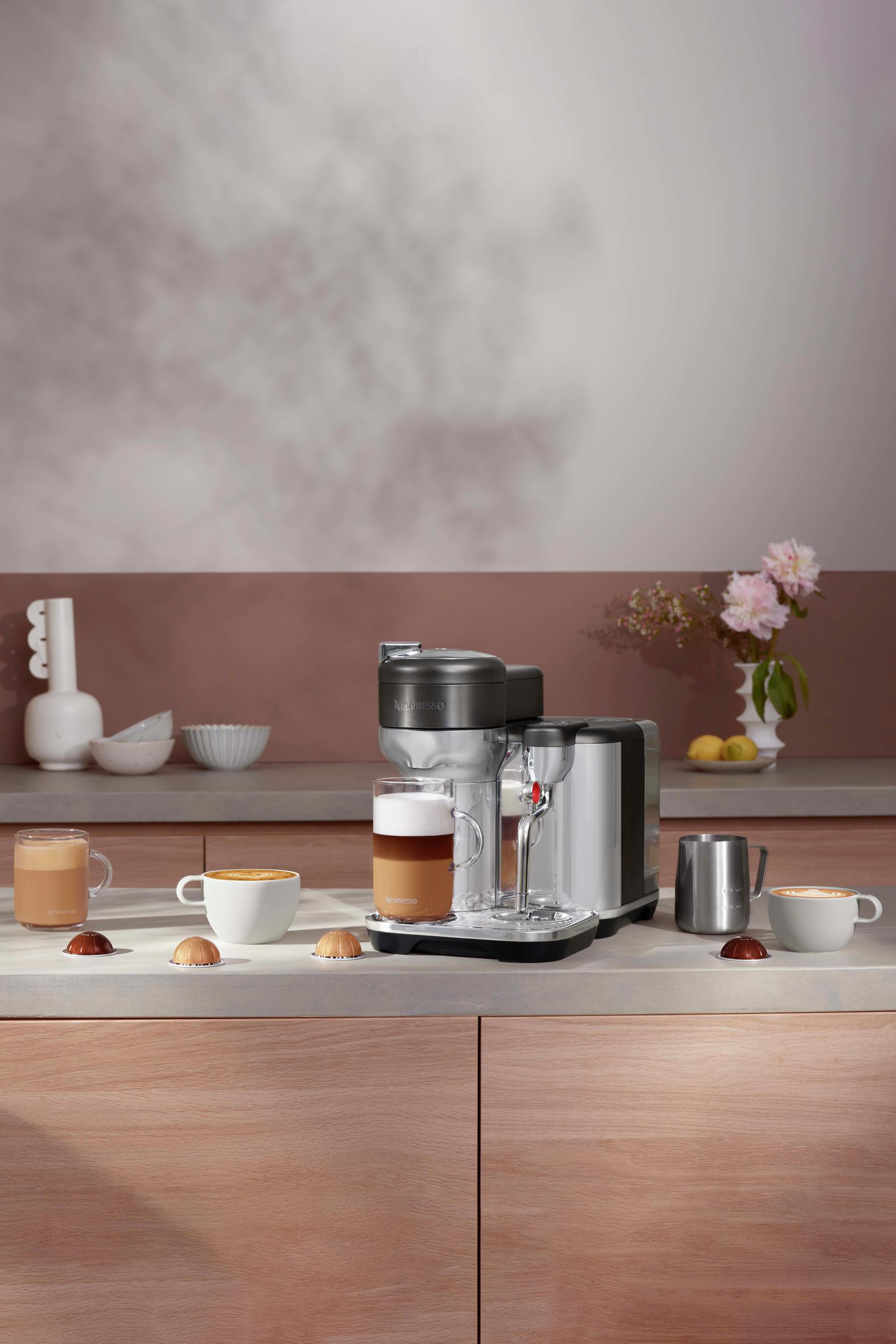 Kaffeemaschine auf der Arbeitsplatte, mit Kaffeetassen und Kapseln. Blumen im Hintergrund. Helles Farbschema.