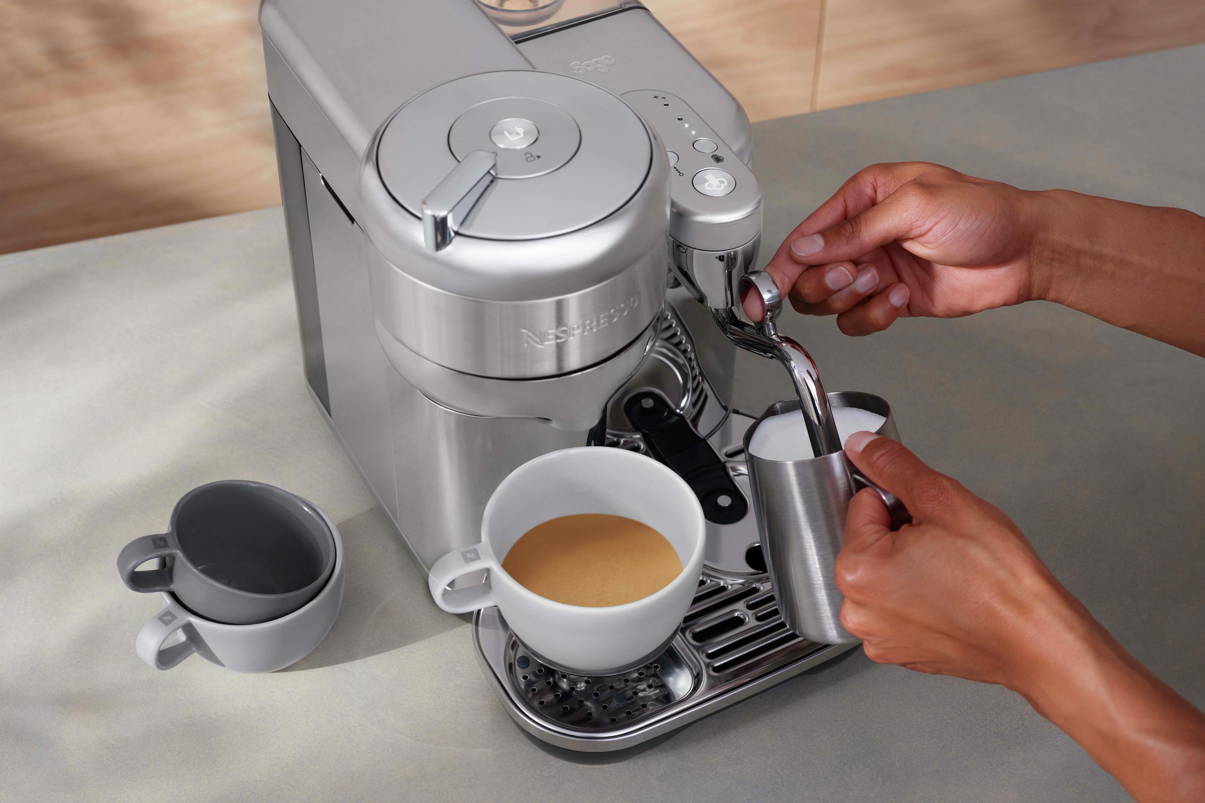 Person bedient silberne Espressomaschine, mit Tassen und Milchaufschäumer.