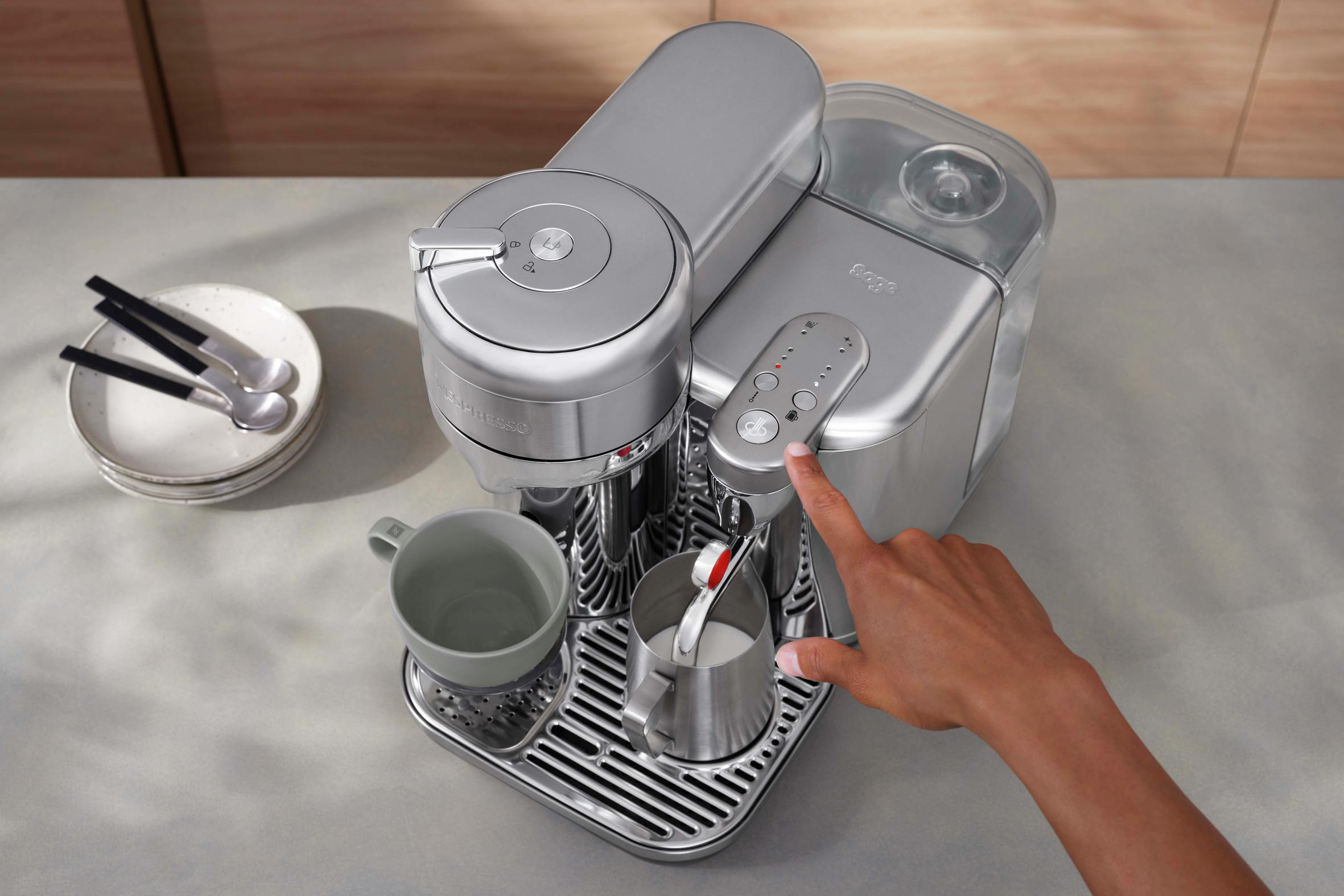 Silberne Nespresso Kaffeemaschine. Eine Hand drückt einen Knopf. Tasse und Zubehör in der Nähe.