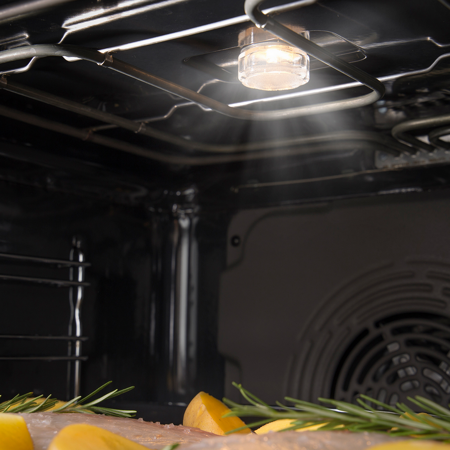 Inside an oven, light shines on food. Rosemary and potatoes sit below the light.
