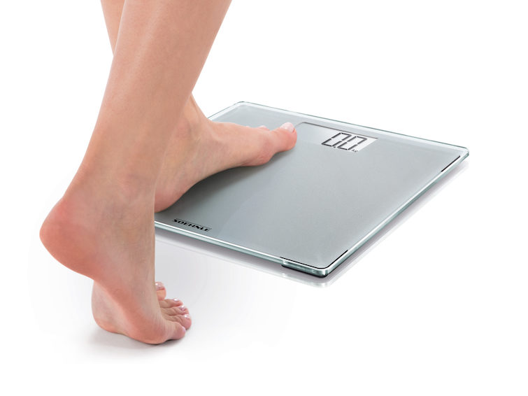 Egy személy digitális mérlegen áll. A mérleg 0,0 kg-ot mutat, fehér háttéren.