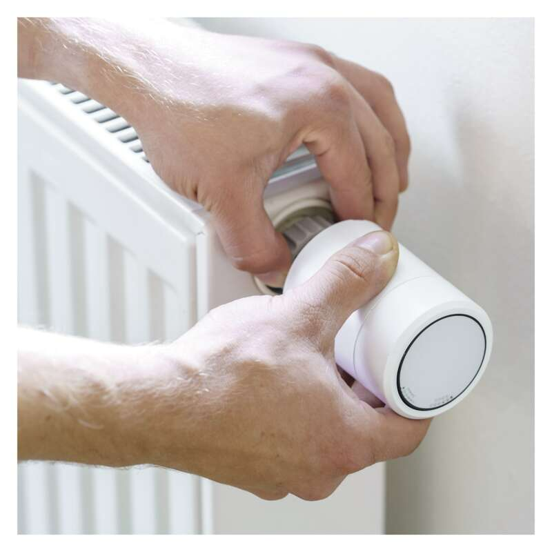 Kéz egy fehér okos termosztátot állít egy radiátoron. A termosztát kör alakú, forgatógombbal.