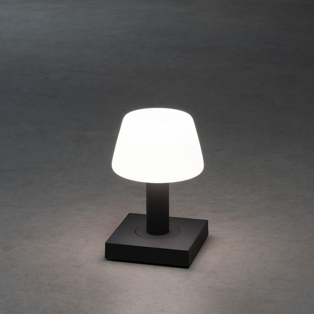 Eine weiß leuchtende Lampe auf schwarzem Sockel. Die Lampe steht auf einer grauen Oberfläche.