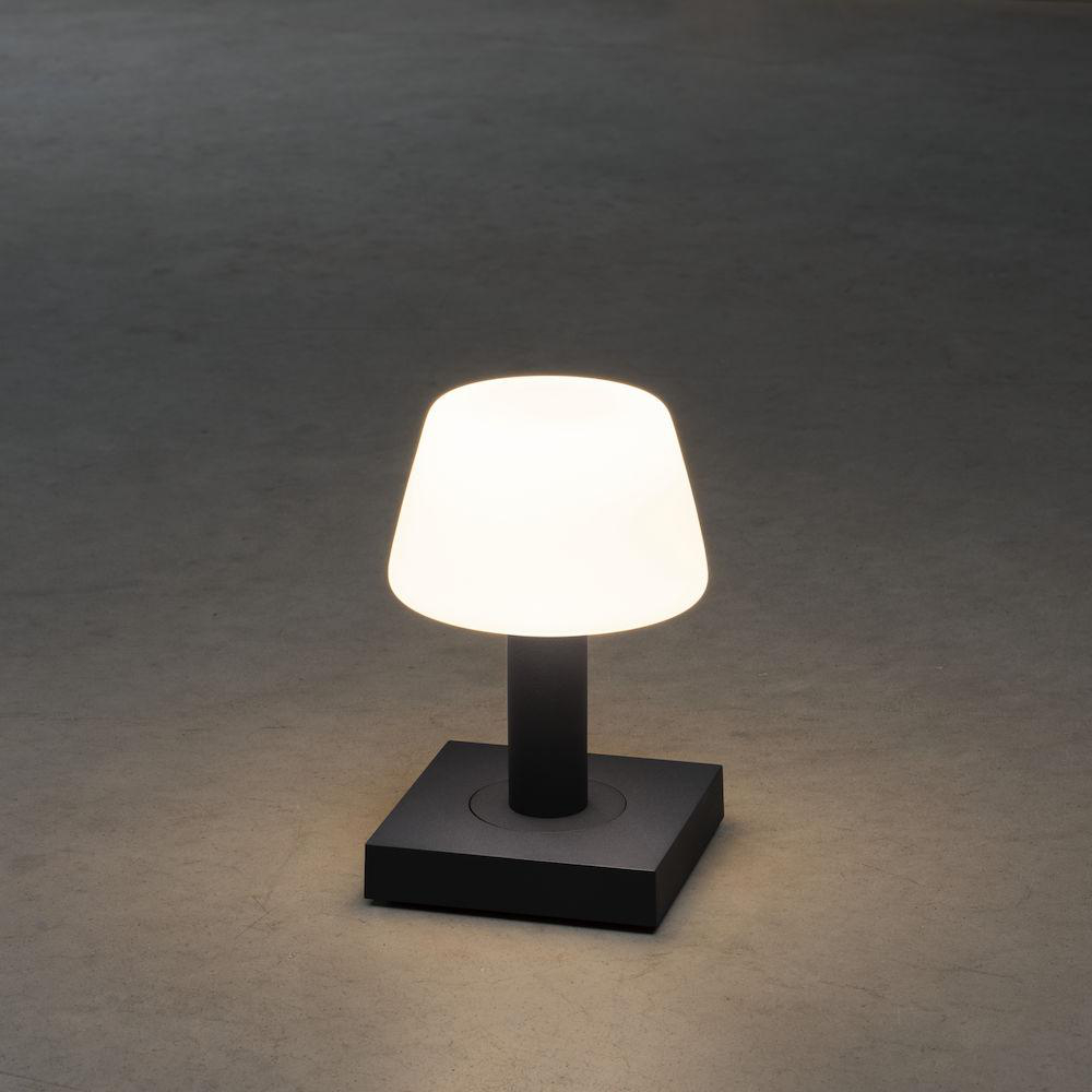 Eine beleuchtete, kleine Tischlampe mit weißem Lampenschirm und dunklem Sockel.