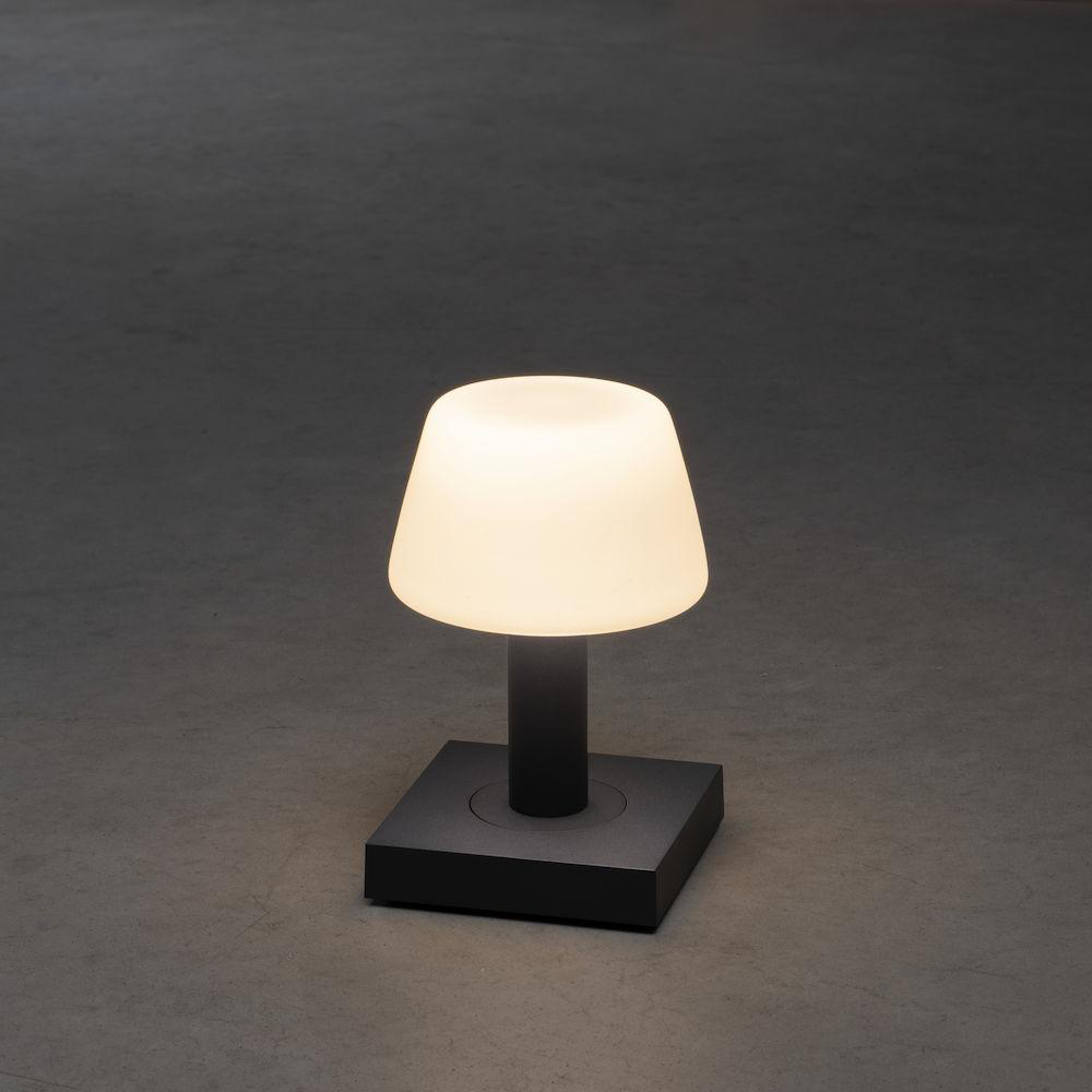 Eine kleine, moderne Lampe mit weißem Schirm auf schwarzem Sockel, vor grauem Hintergrund.