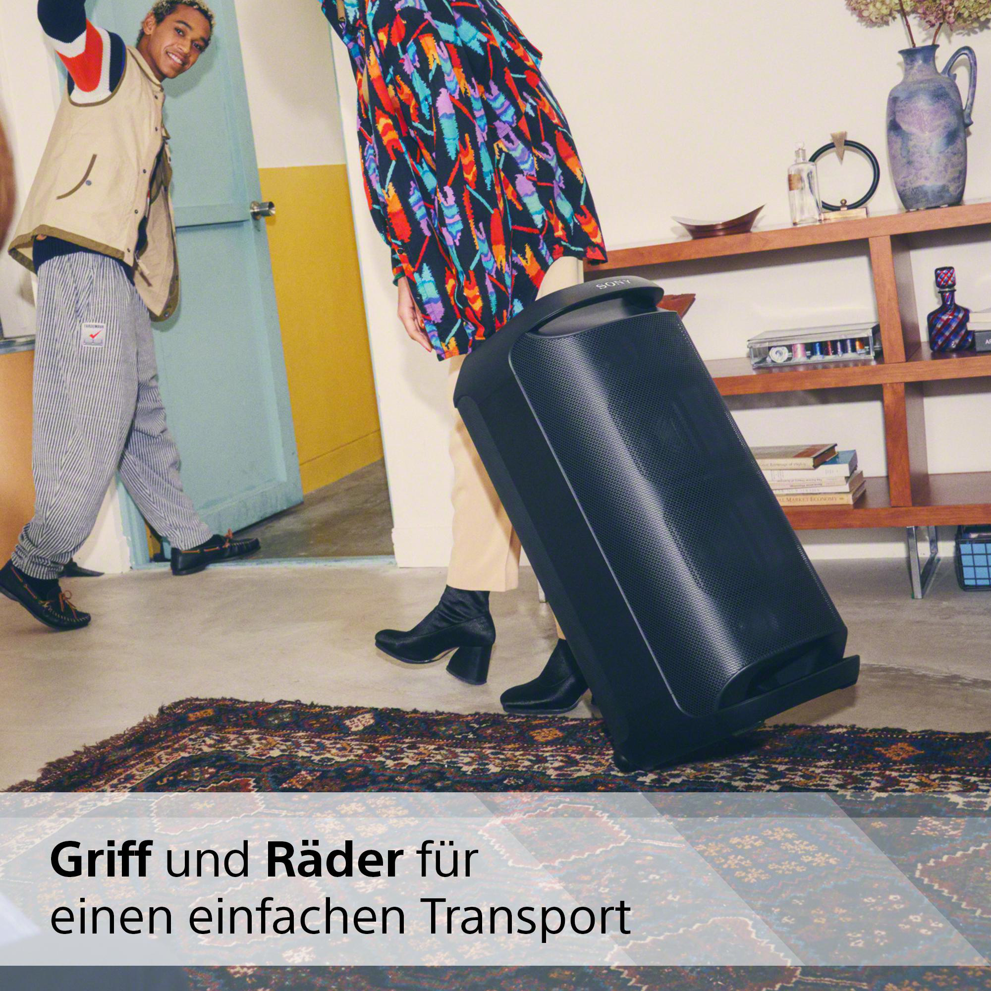 Zwei Personen tragen einen schwarzen Lautsprecher mit Rädern auf einem Teppich.