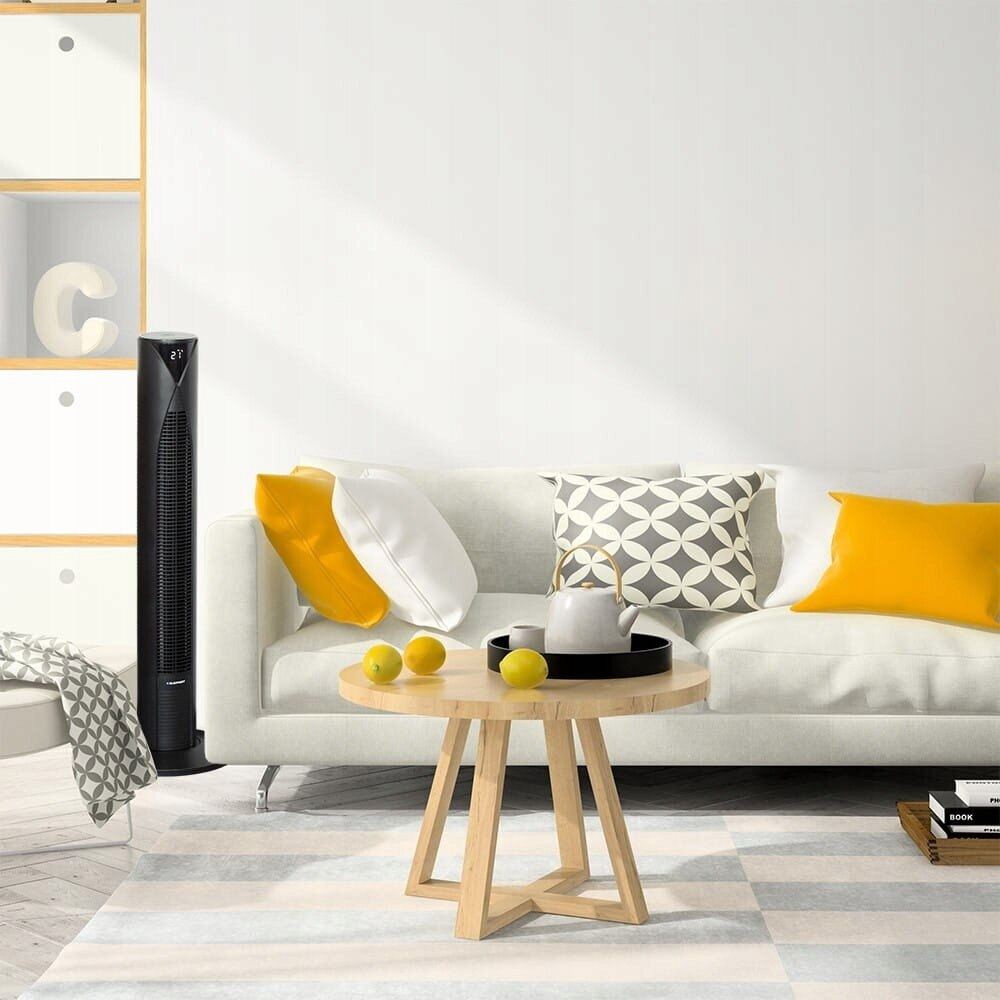 Salon z białą sofą, żółtymi poduszkami, okrągłym drewnianym stołem, czarnym wentylatorem i białą ścianą.