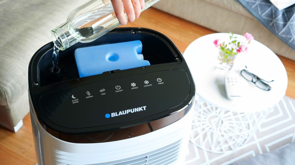 Ręka nalewa wodę do klimatyzatora Blaupunkt. W środku niebieski wkład chłodzący. Blisko stół z kwiatami, okularami i pilotem.